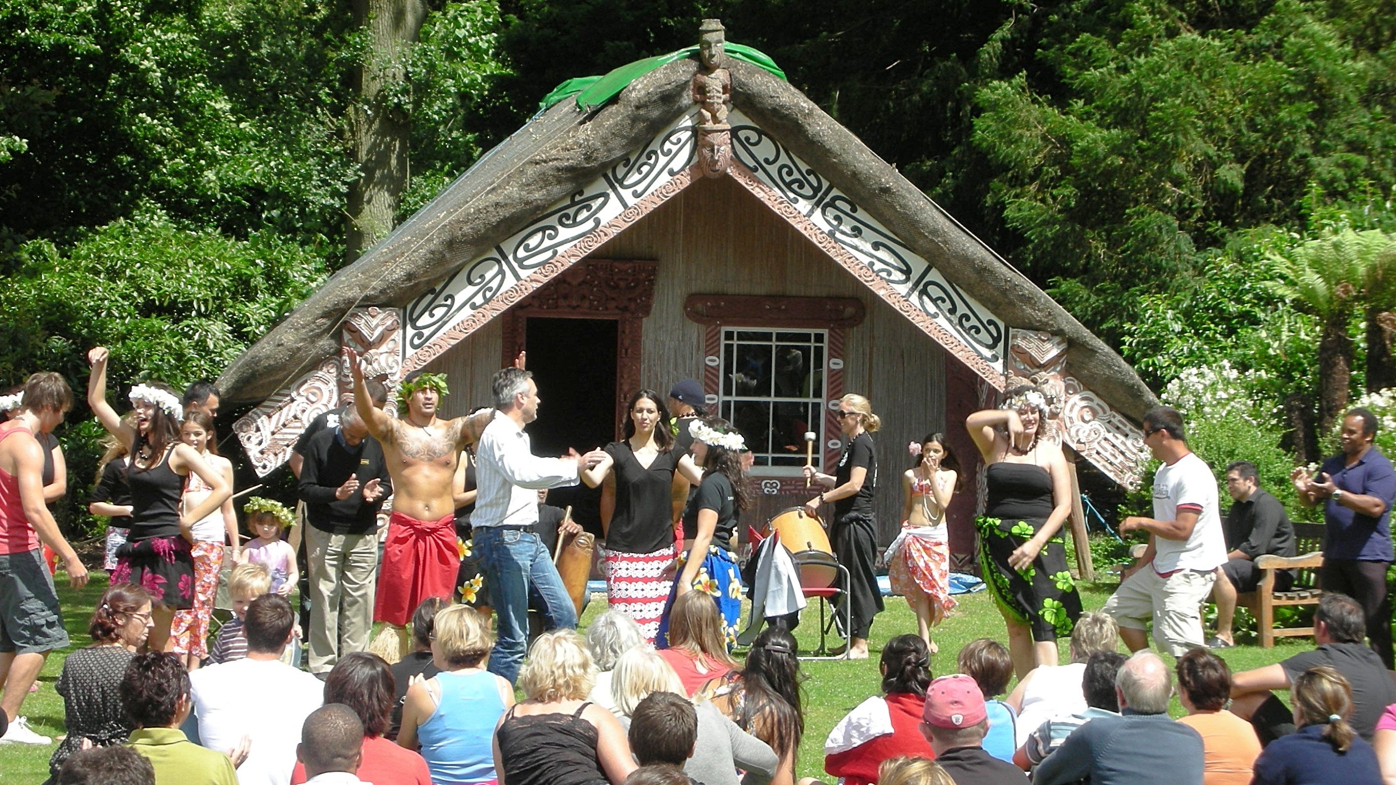 A group of people performing traditional Māori dance in front of a Māori meeting house with an audience sitting on the ground watching