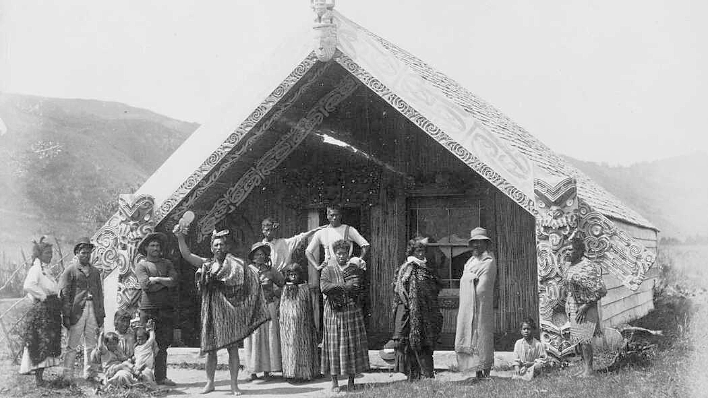 A black and white image of a Maori wharenui - a wooden meeting house decorated with carvings - with Maori people standing outside.