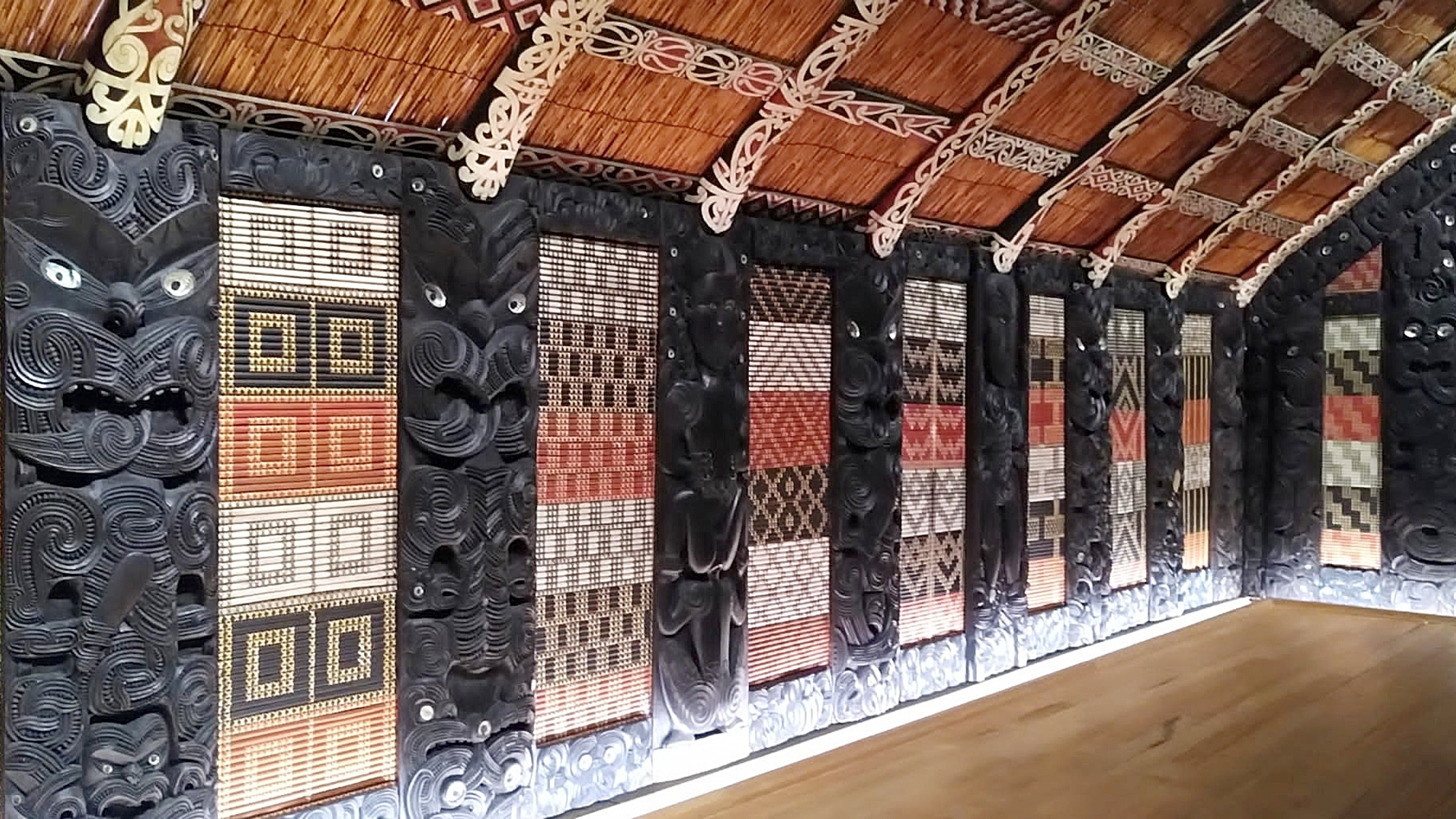 The interior of Te Rauru, a Māori wharenui (meeting house) lined with black-painted carvings interspersed with red, white and black woven panels