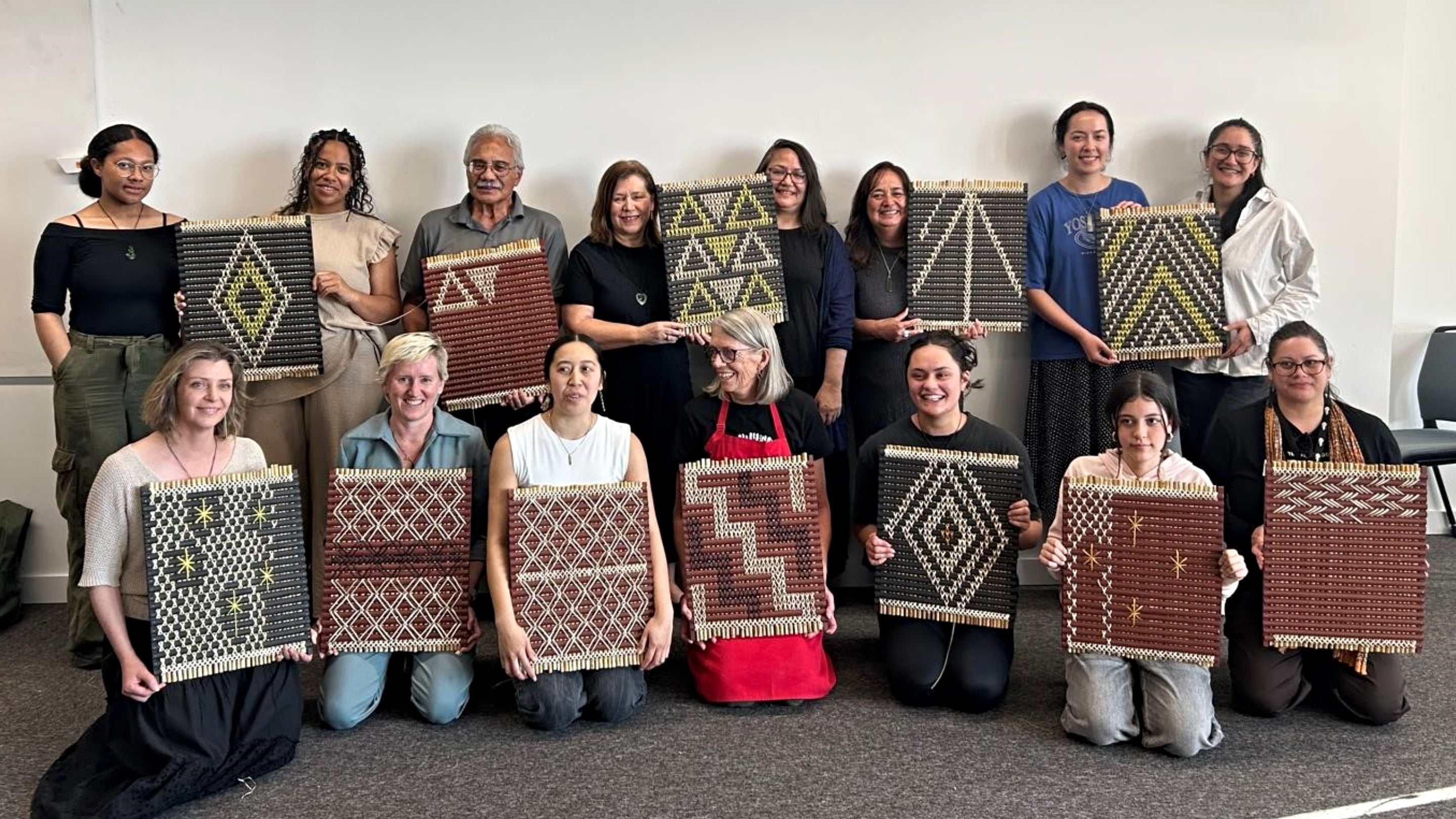 Two rows of people, one standing behind, one kneeling in front, holding up woven tukutuku panels