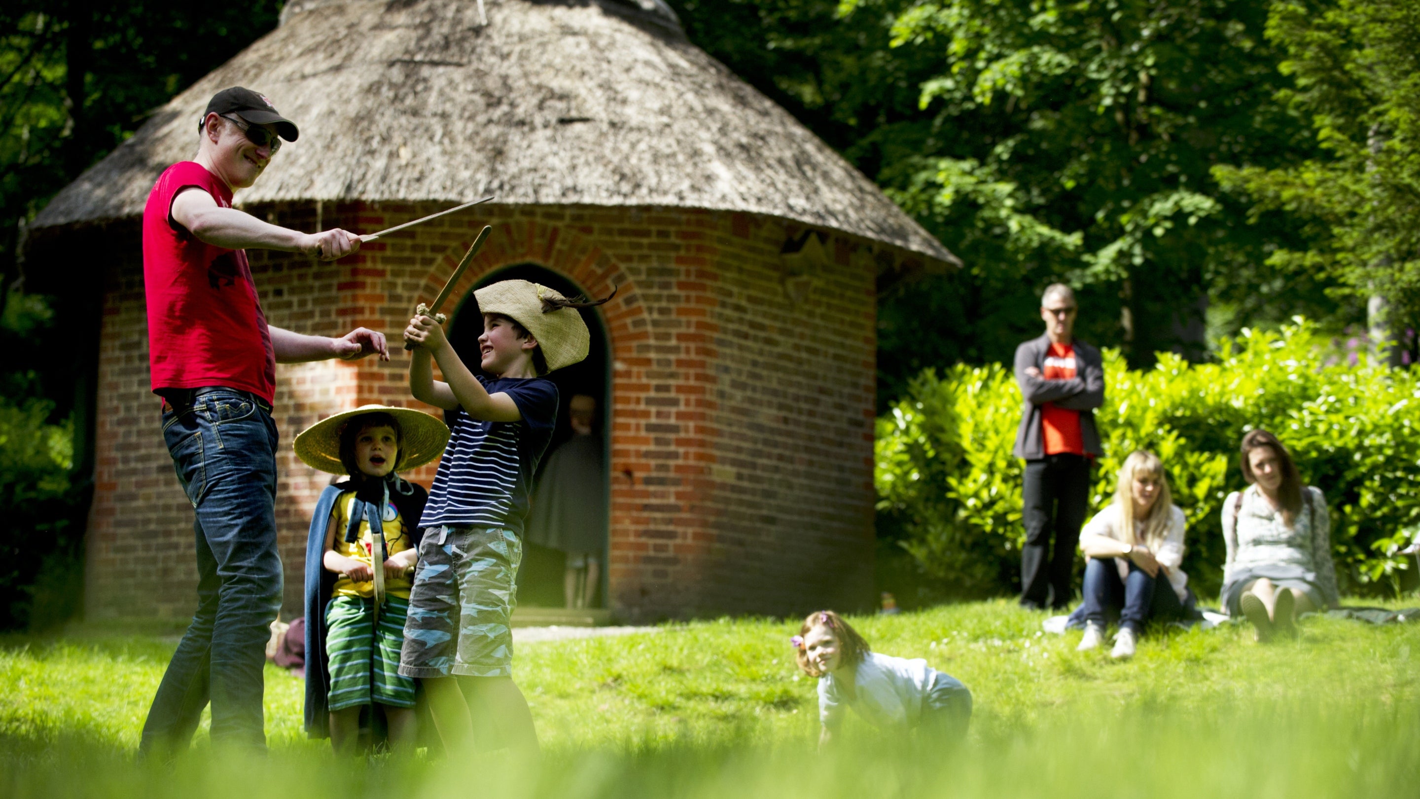 Father and son having an pretend sword fight with smaller sister watching in front of the Thatched cottage at Claremont Landscape Garden