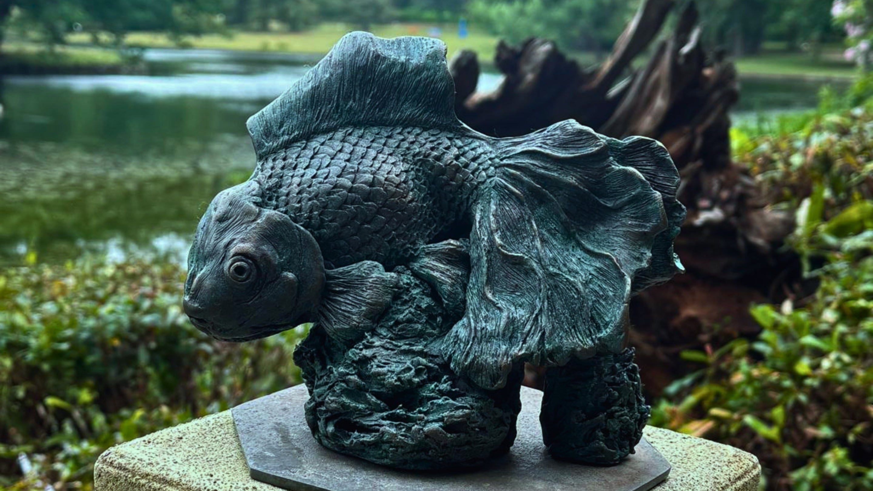 The image shows a sculptural artwork of a fish displayed outdoors in a landscaped garden setting. The fish has a rounded body with pronounced, textured scales and flowing fins.