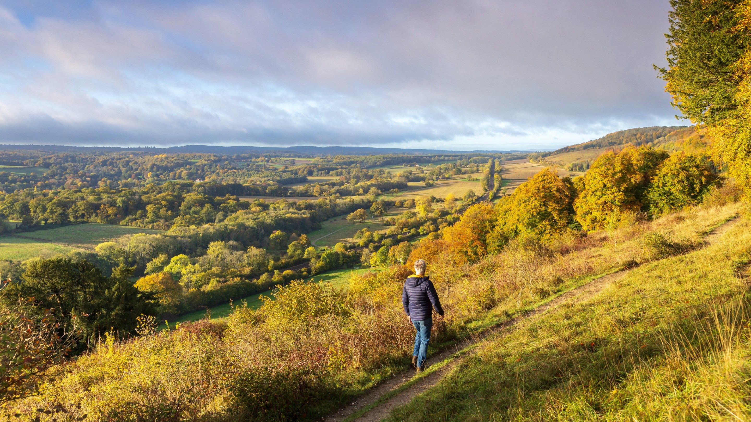 A man in a blue jacket and jeans walking along the path at the top of the hillside in autumn, with a view of the valley below
