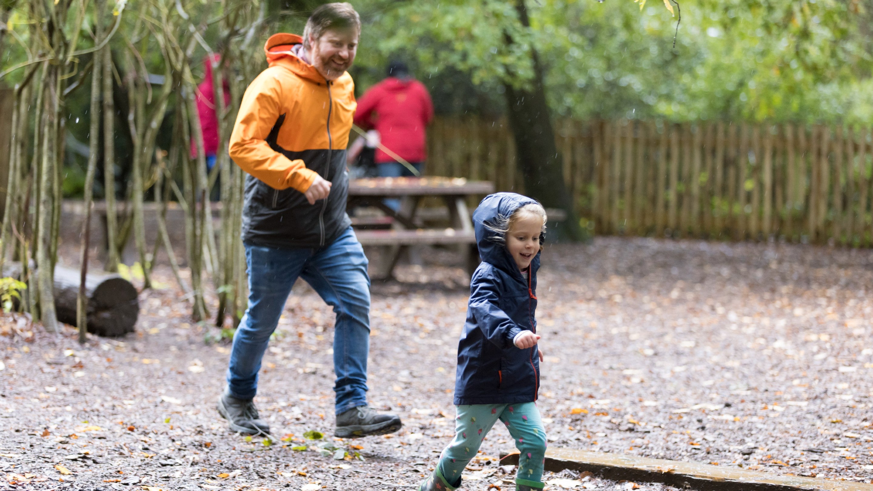 Father running with his daughter in the natural play area with a picnic bench and willow tunnel in the background.