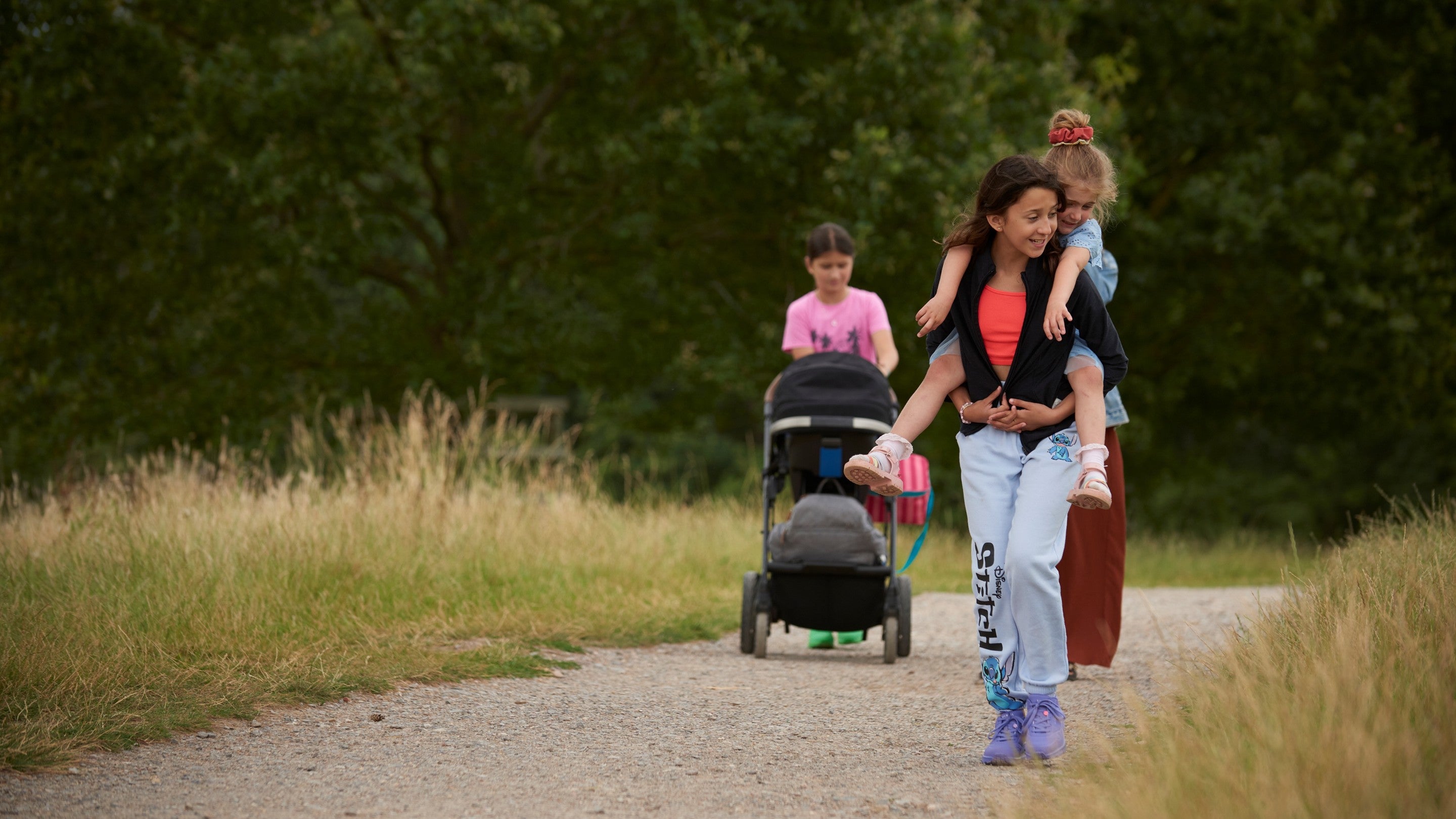 Two children walk along a gravel path through tall grass, with one child carrying a smaller child on their back. Behind them, another child pushes a stroller along the same path. Trees and greenery line the route.