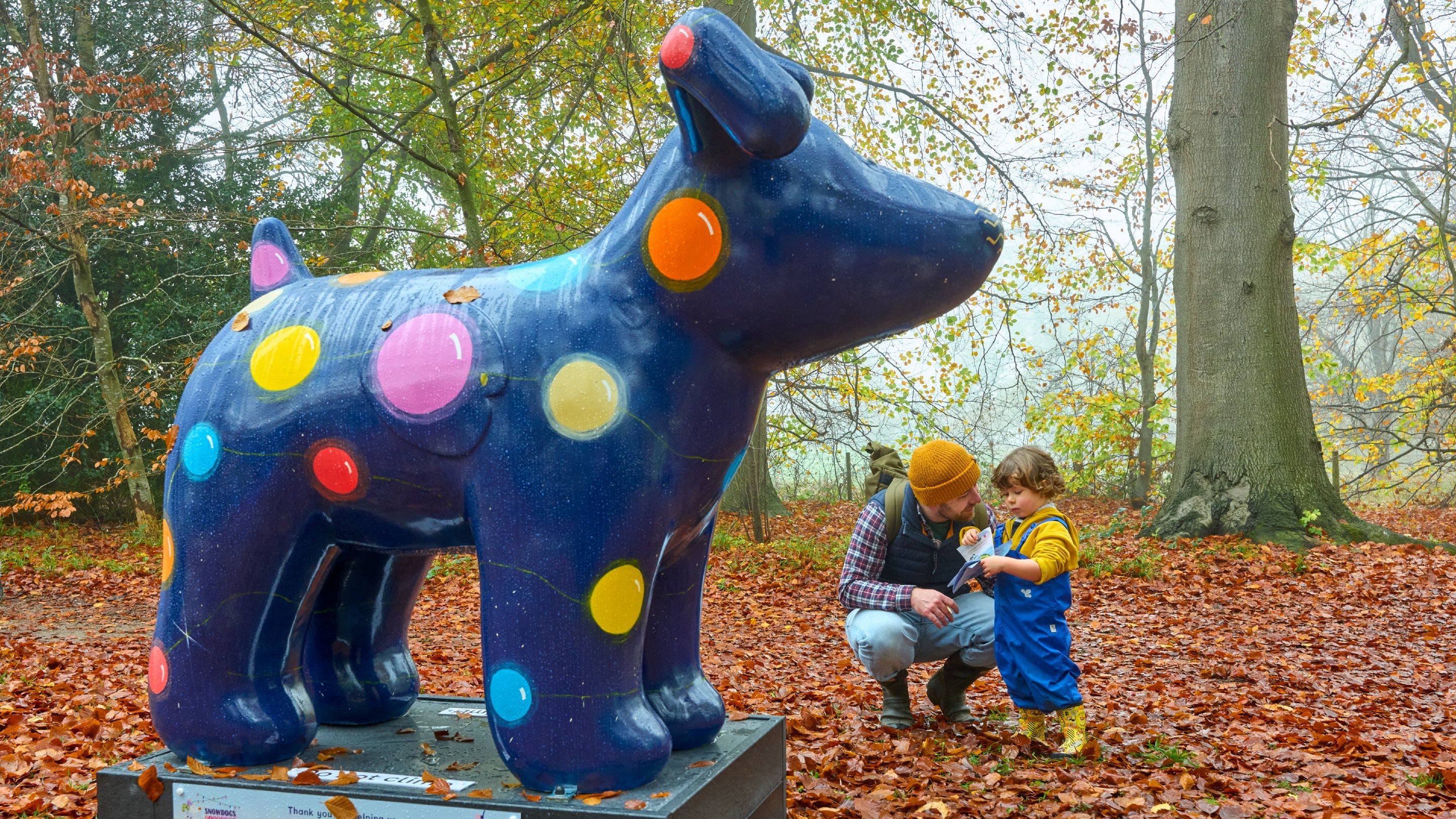 A man crouched down next to a toddler, both in woolly hats, looking at a sculpture of a dark blue Snowdog painted with large brightly coloured circles resembling lights.