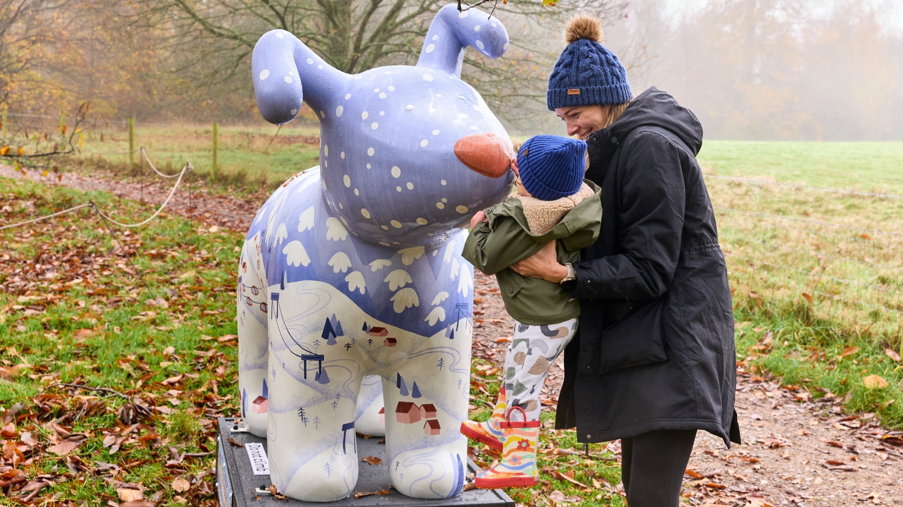 Woman bobble hat lifting up a toddler to touch the nose of a large snow dog sculpture, which is decorated with an alpine ski view.