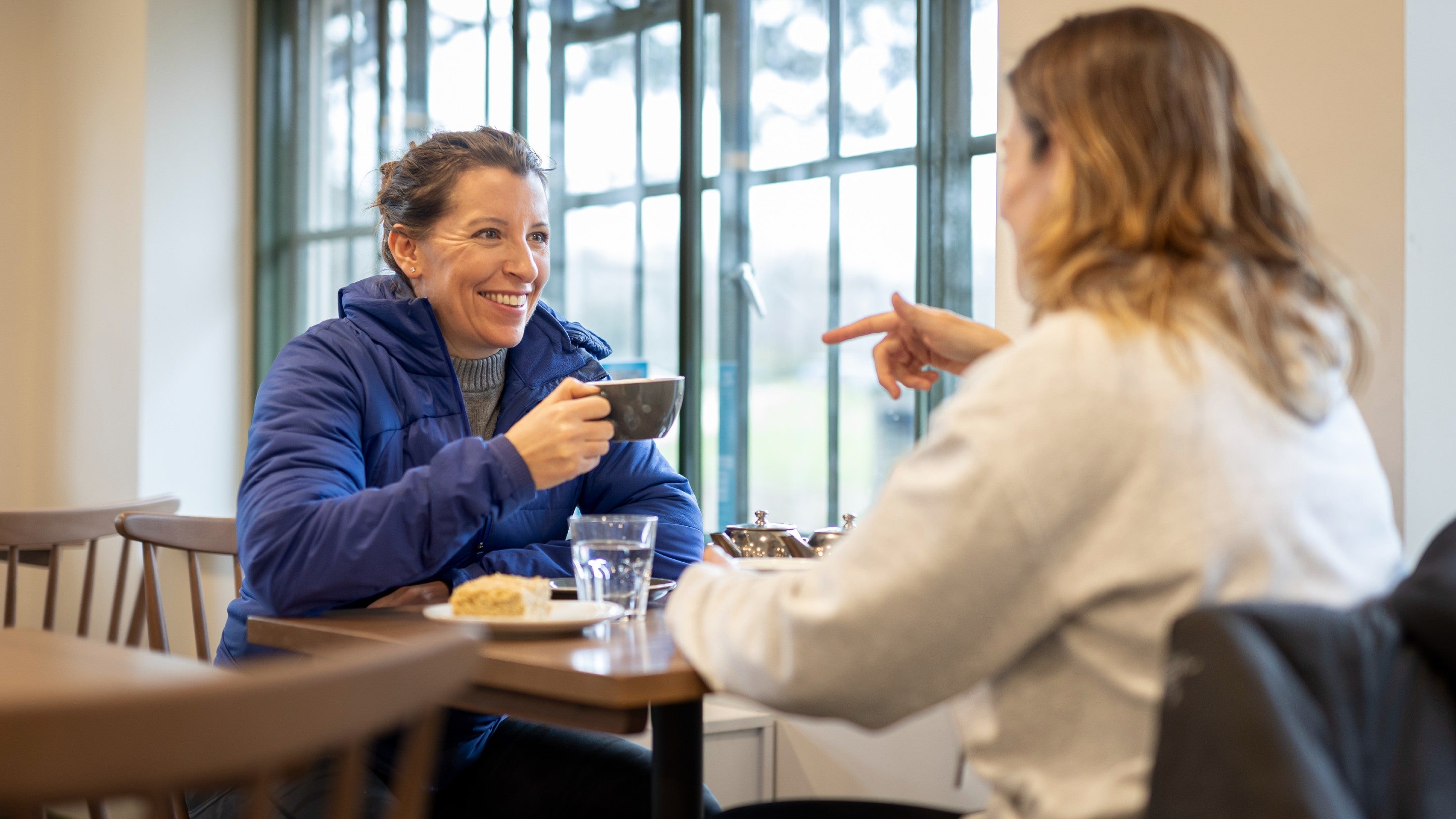 Two women sitting opposite each other at a table, smiling, with cups of tea and a flapjack on the table