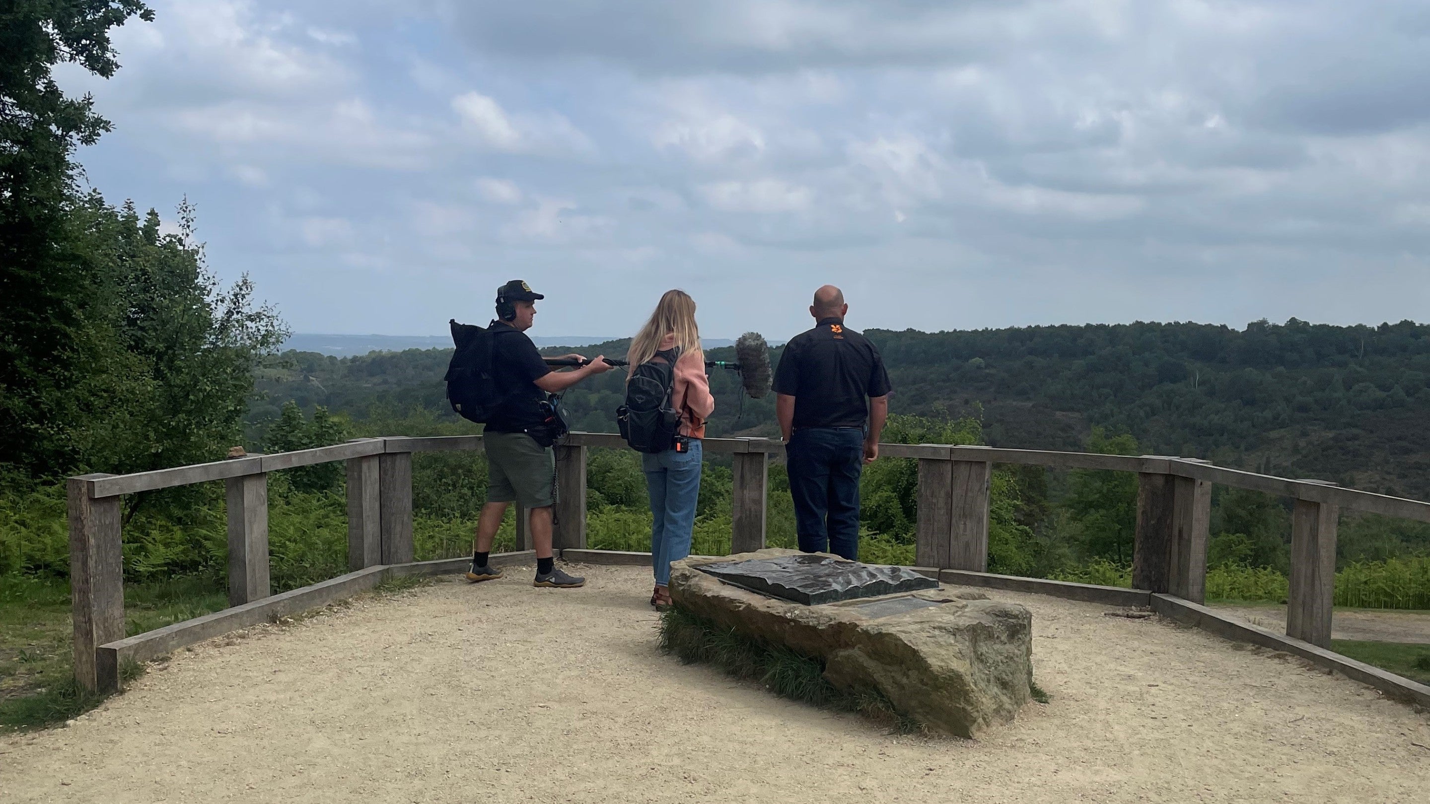 Three people with their backs to the camera enjoying the view at Hindhead Commons. The ranger is being interviewed by a woman in a pink jacket and a sound recordist is standing next to her.