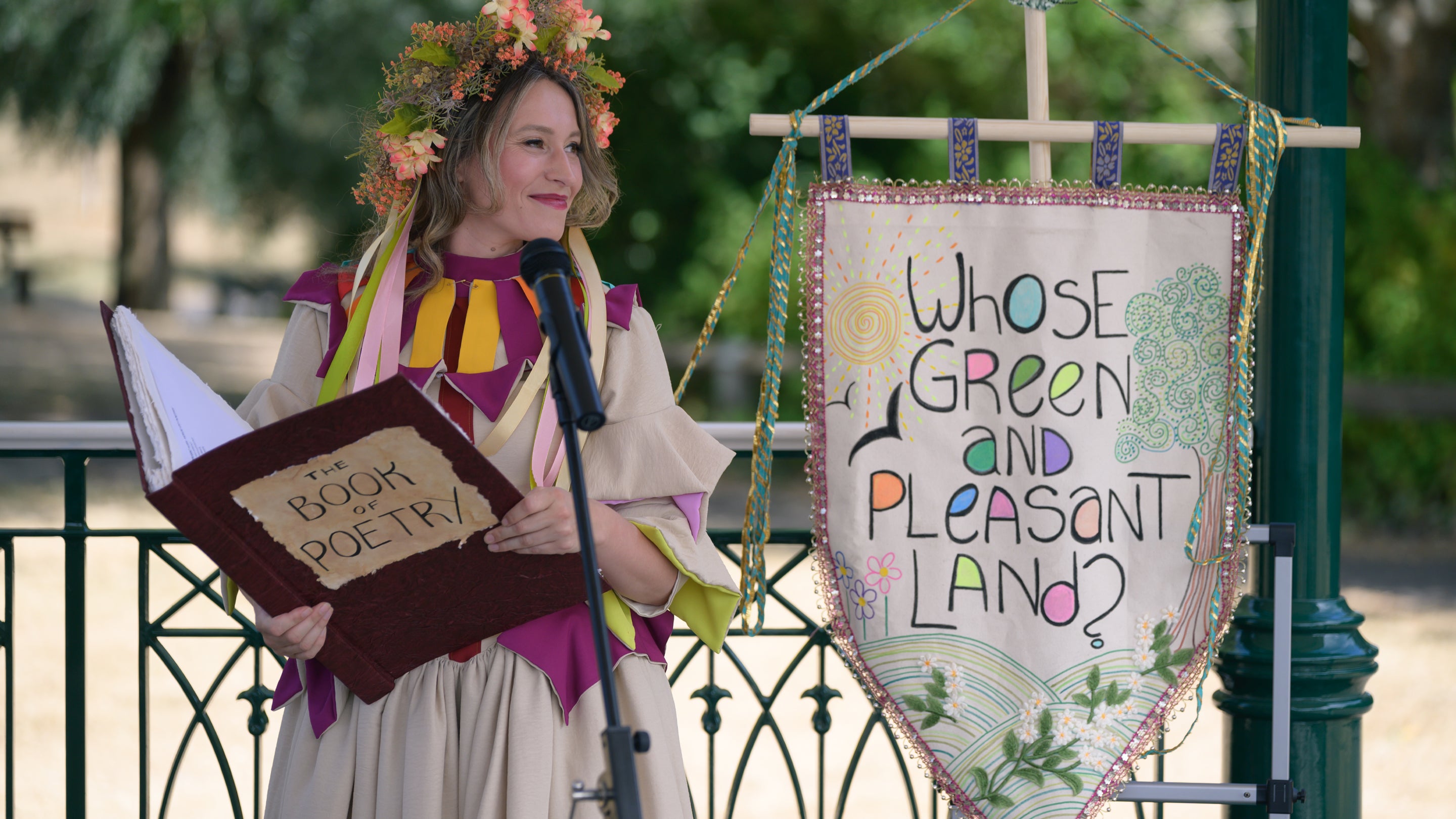 Woman in a cream and red dress and a head garland, reading from a large red poetry book, standing next to a cloth banner embroidered with the words: Whose green and pleasant land?