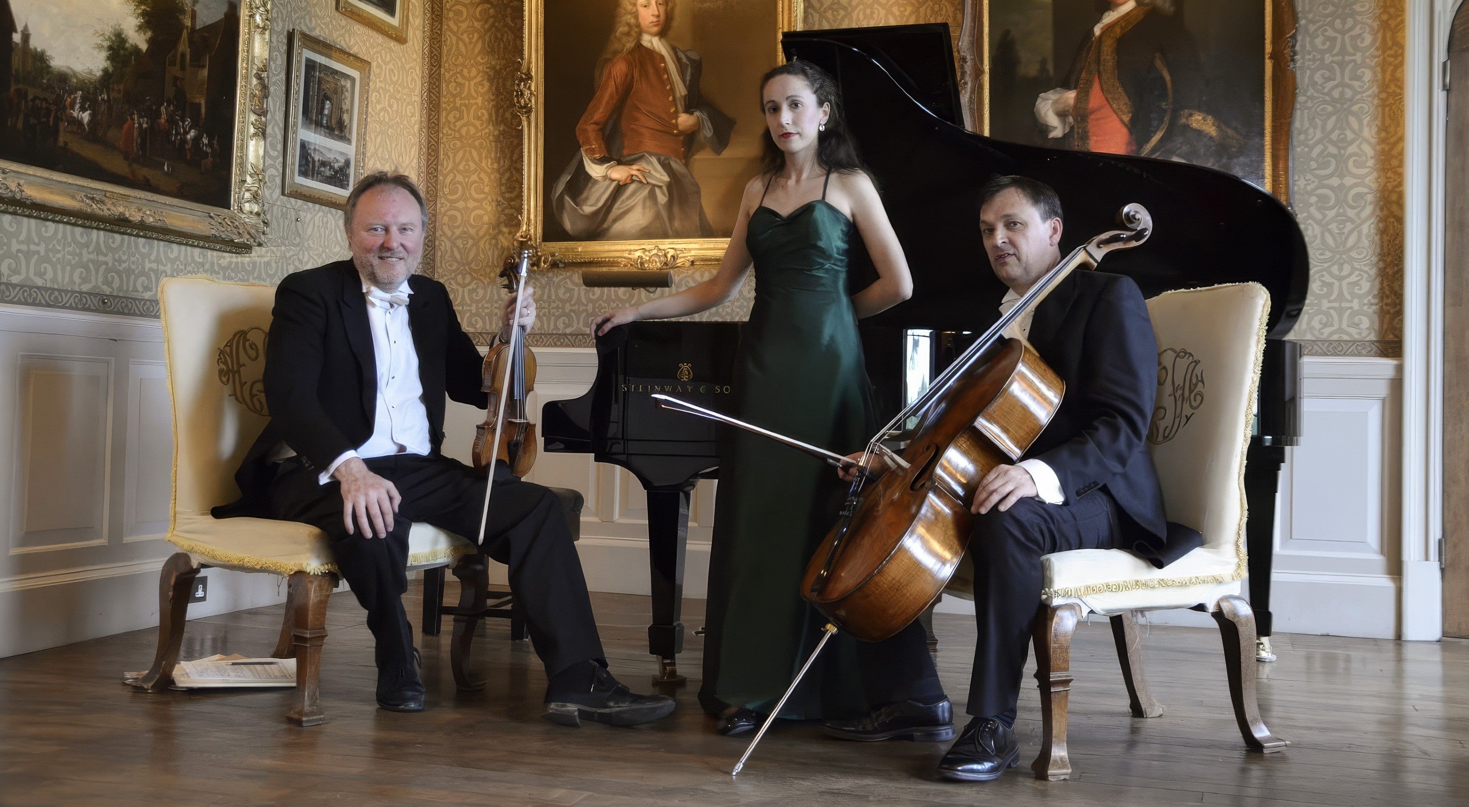 A woman in a green evening dress standing in front of a grand piano. Two men sit either side of her, in evening dress, one holds a cello and the other holds a violin. They are in a country house with oil paintings hanging on the wall behind them.