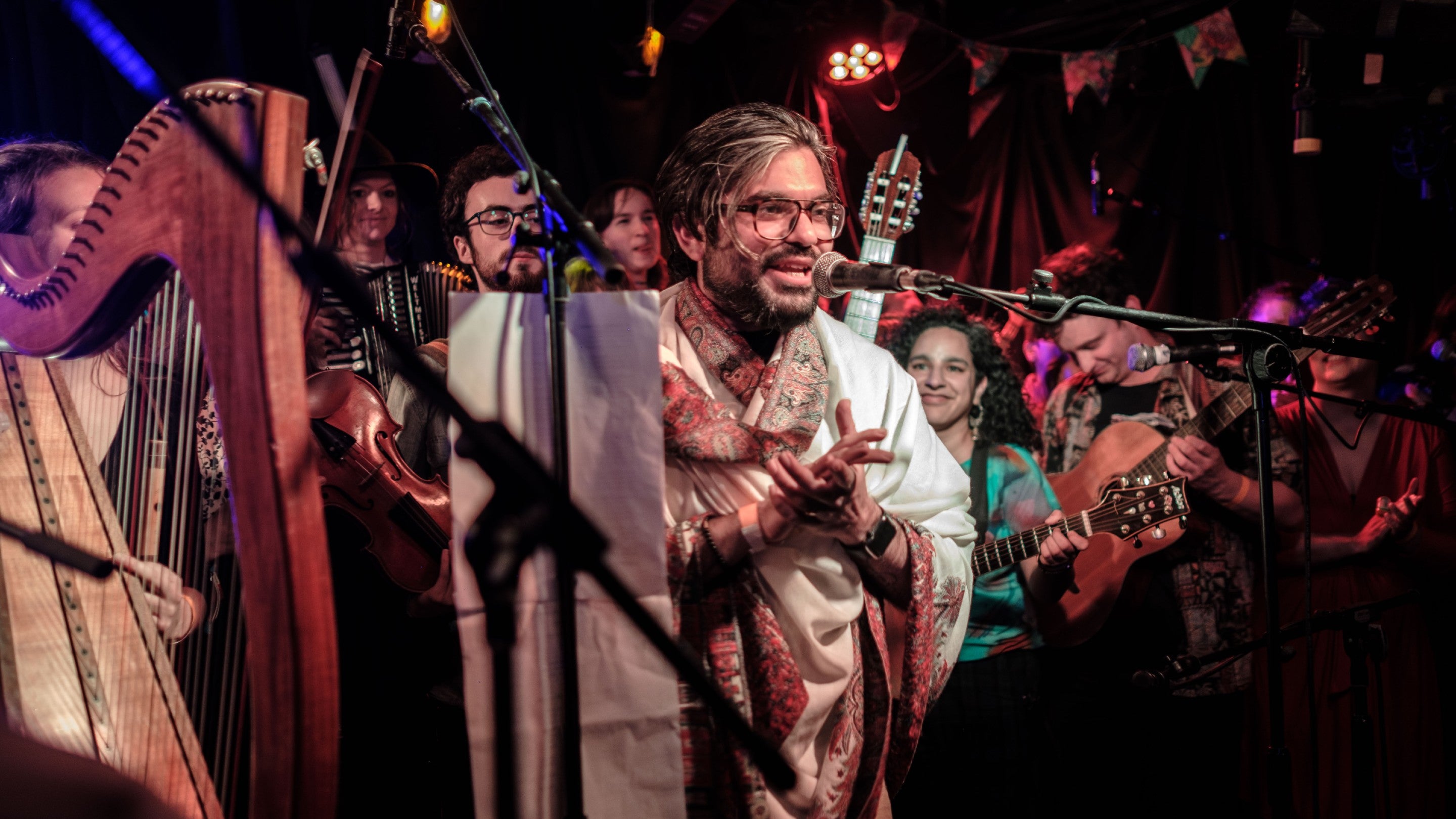 Man in a colourful outfit clapping, surrounded by people playing all sorts of instruments including a harp in the foreground