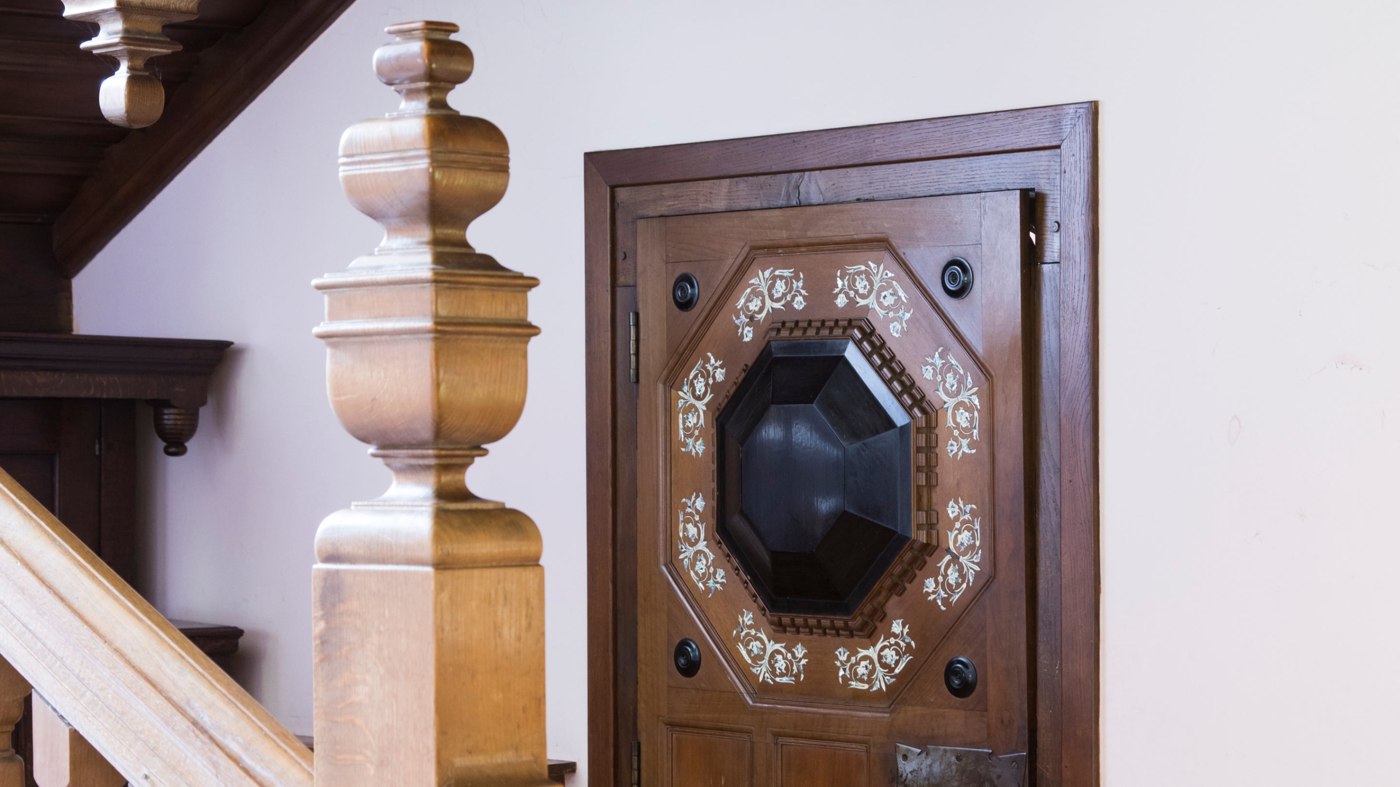 IN the background an oak door is inlayed with fine white bone inlay with mother of pearl.  In the foreground is the carved post of a staircase