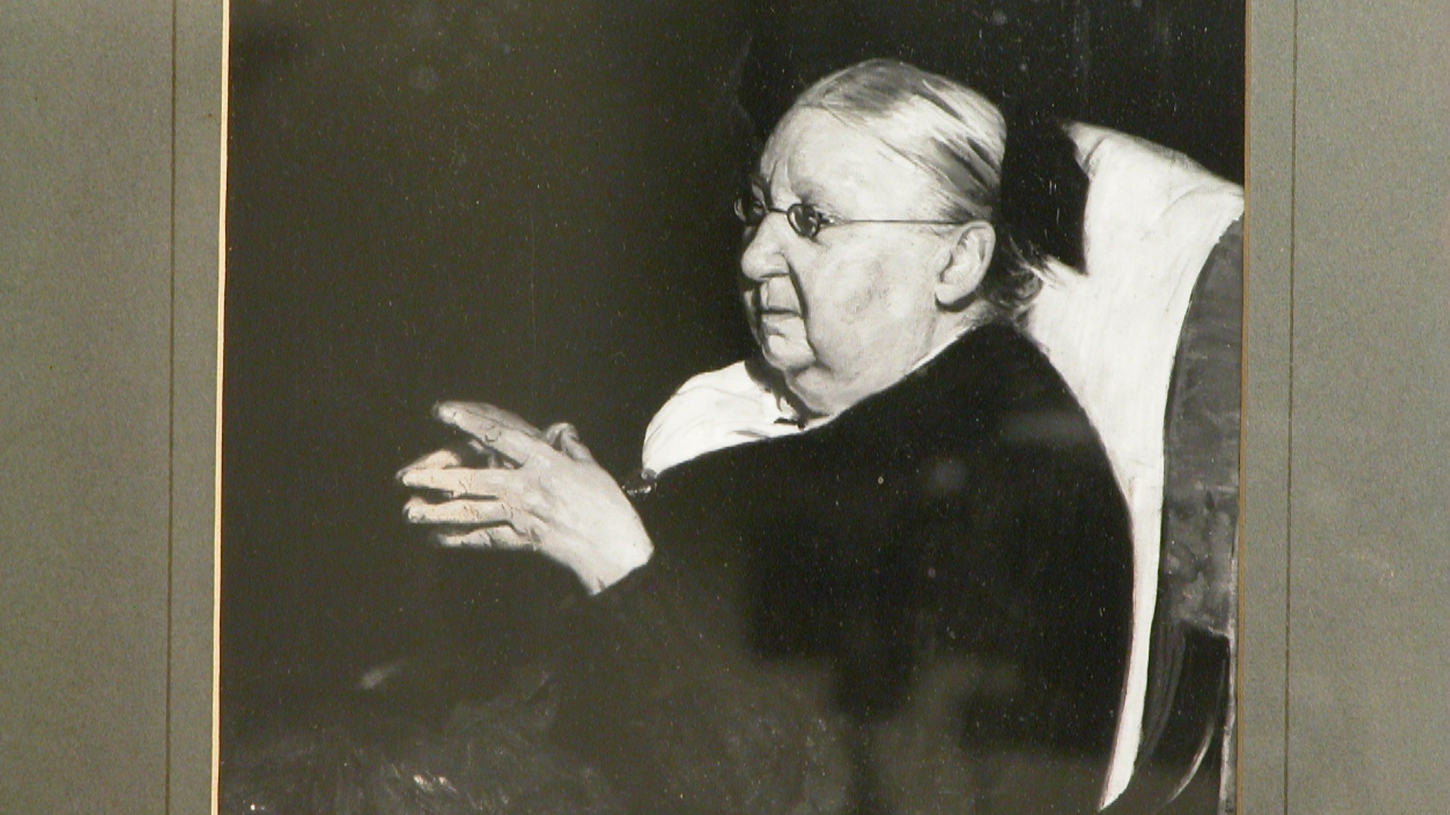black and white photo of a portrait of an elderly woman sitting with her hands clasped , she had round spectacles and her hair tied back with a ribbon.