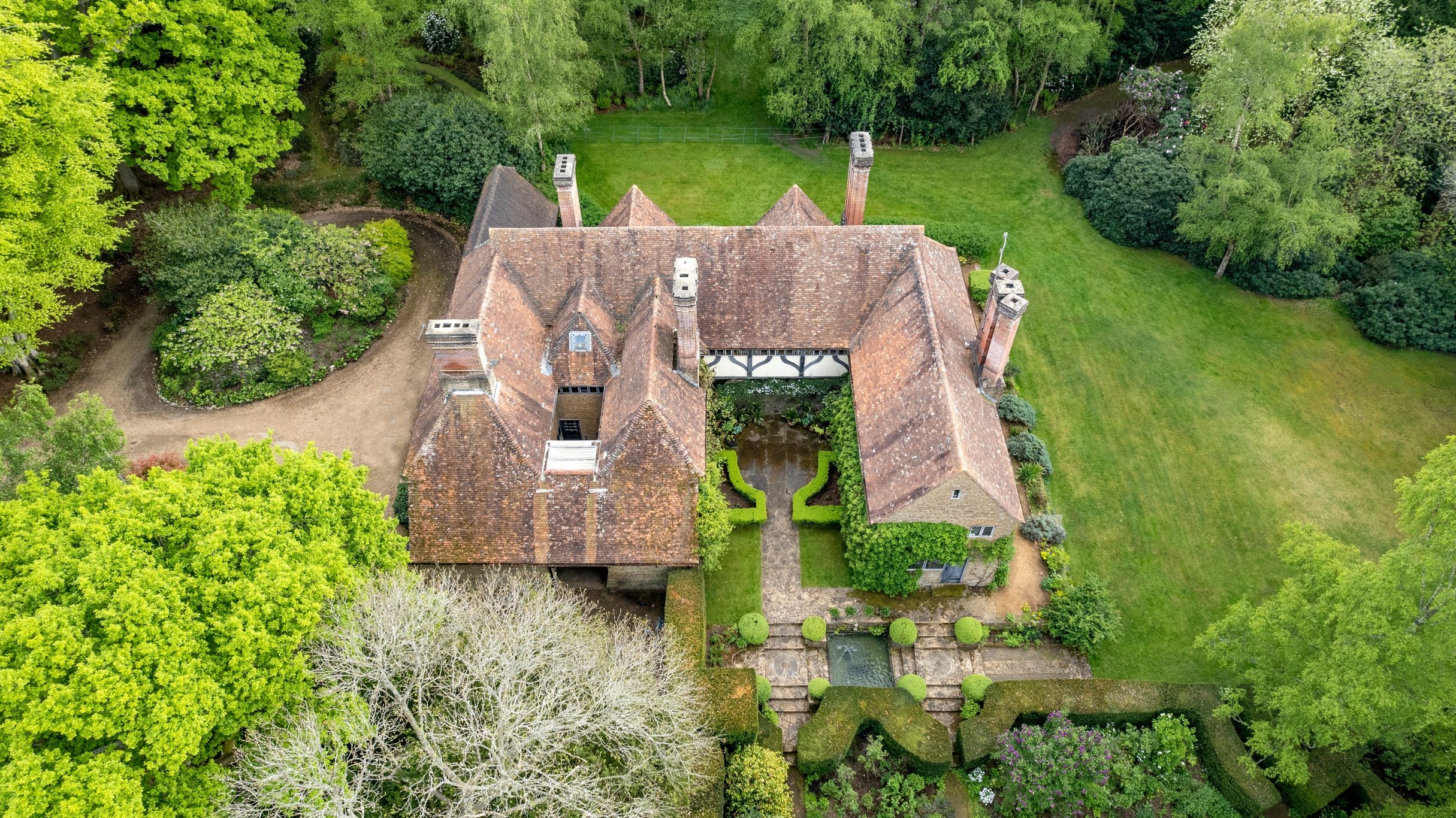 An aerial view of a large red tiled roof on a large house with 5 chimneys surrounded by green lawn and woodland