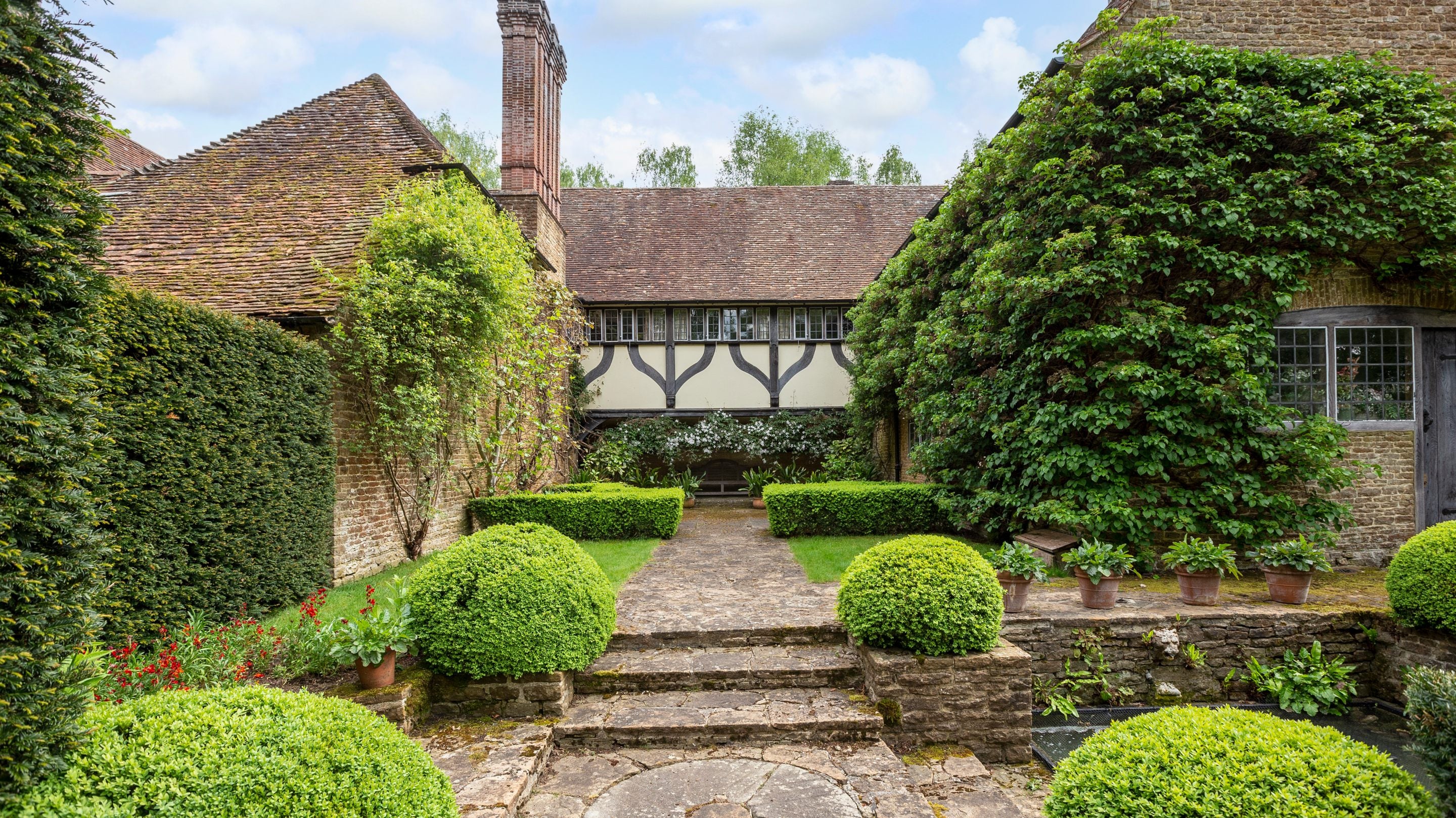 Stone steps lead to a shady courtyard garden laid out in a geometric style with round box spheres a square pond and surrounded by an old house on three sides.