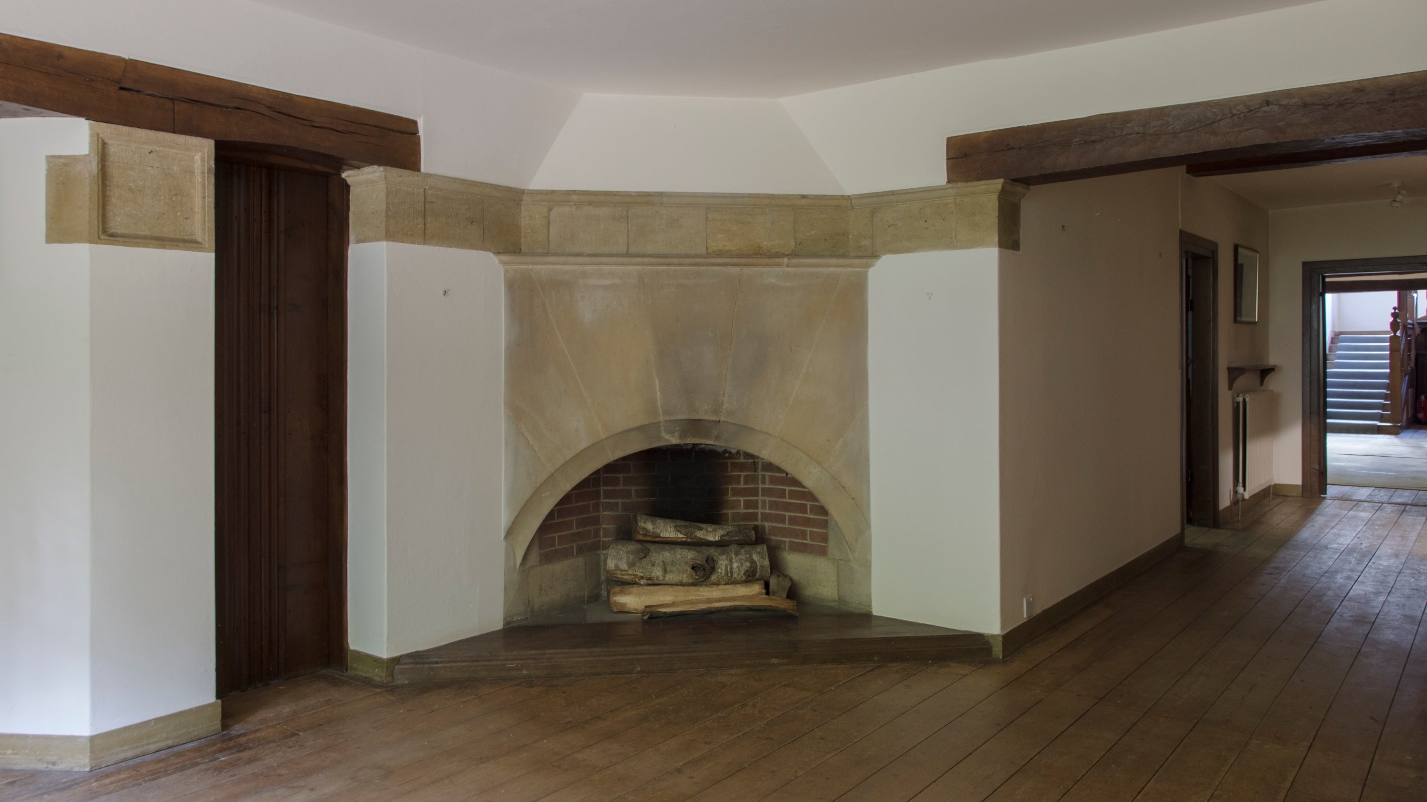 A wooden floor leads to a stone fireplace carved into an arch above wooden logs.  A corridor leads to the right towards an open door and a staircase.  Munstead Wood, Surrey