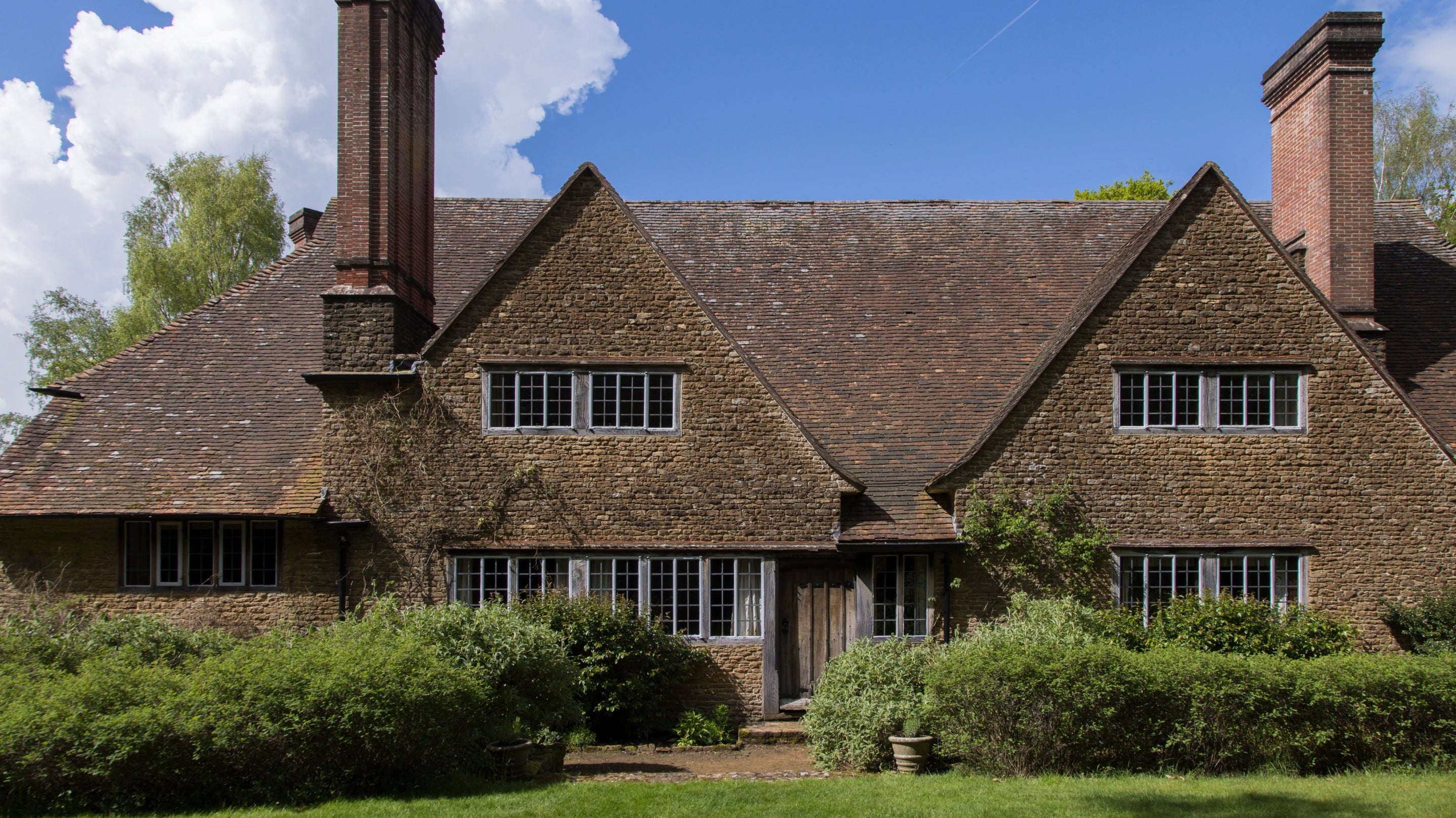 Brick and stone south elevation to the house at Munstead Wood.  The ground floor of the house is partially hidden by bushy shrubs.