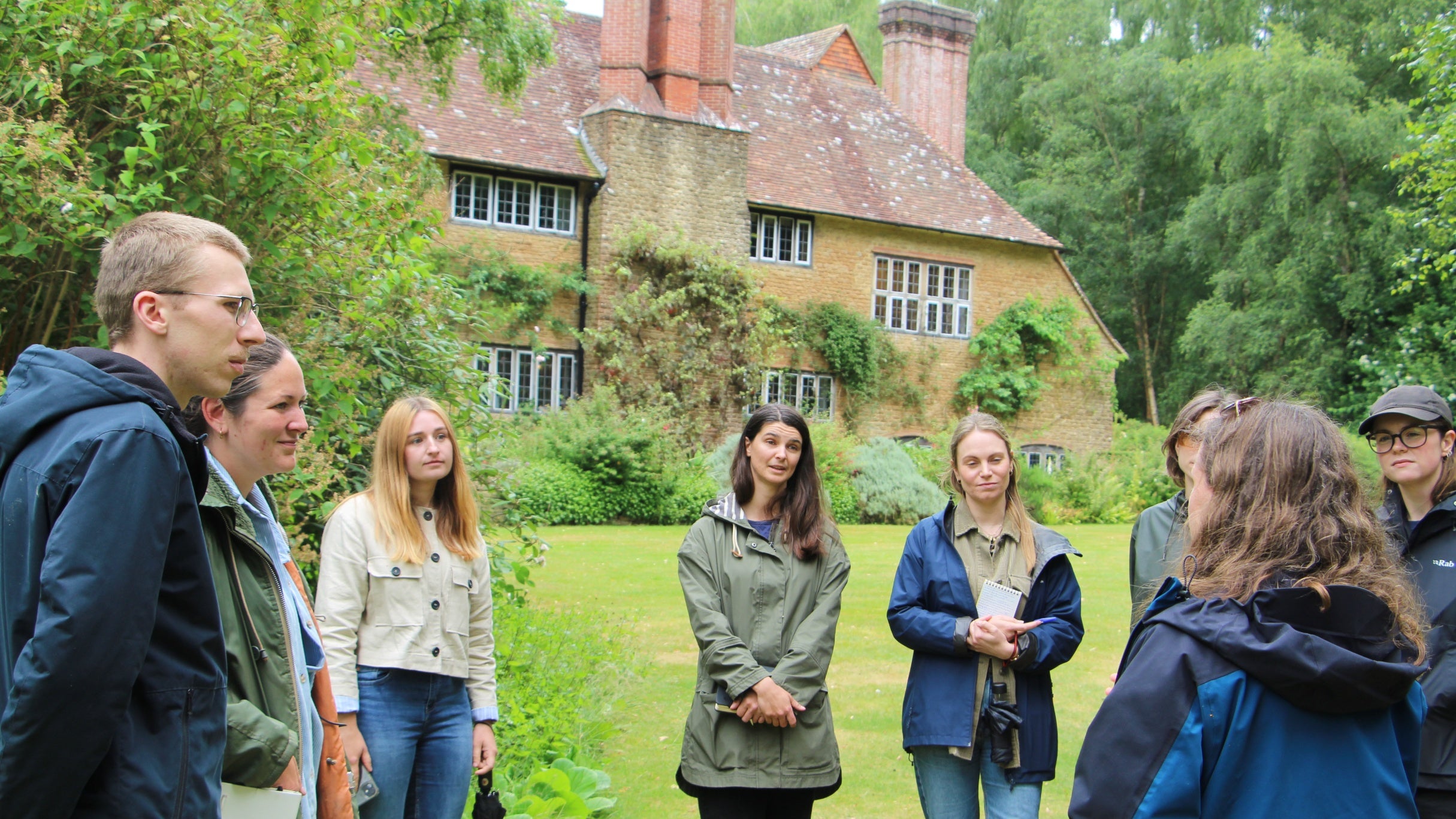 A group of people stand in a circle listening to somebody speak with a large house and garden behind