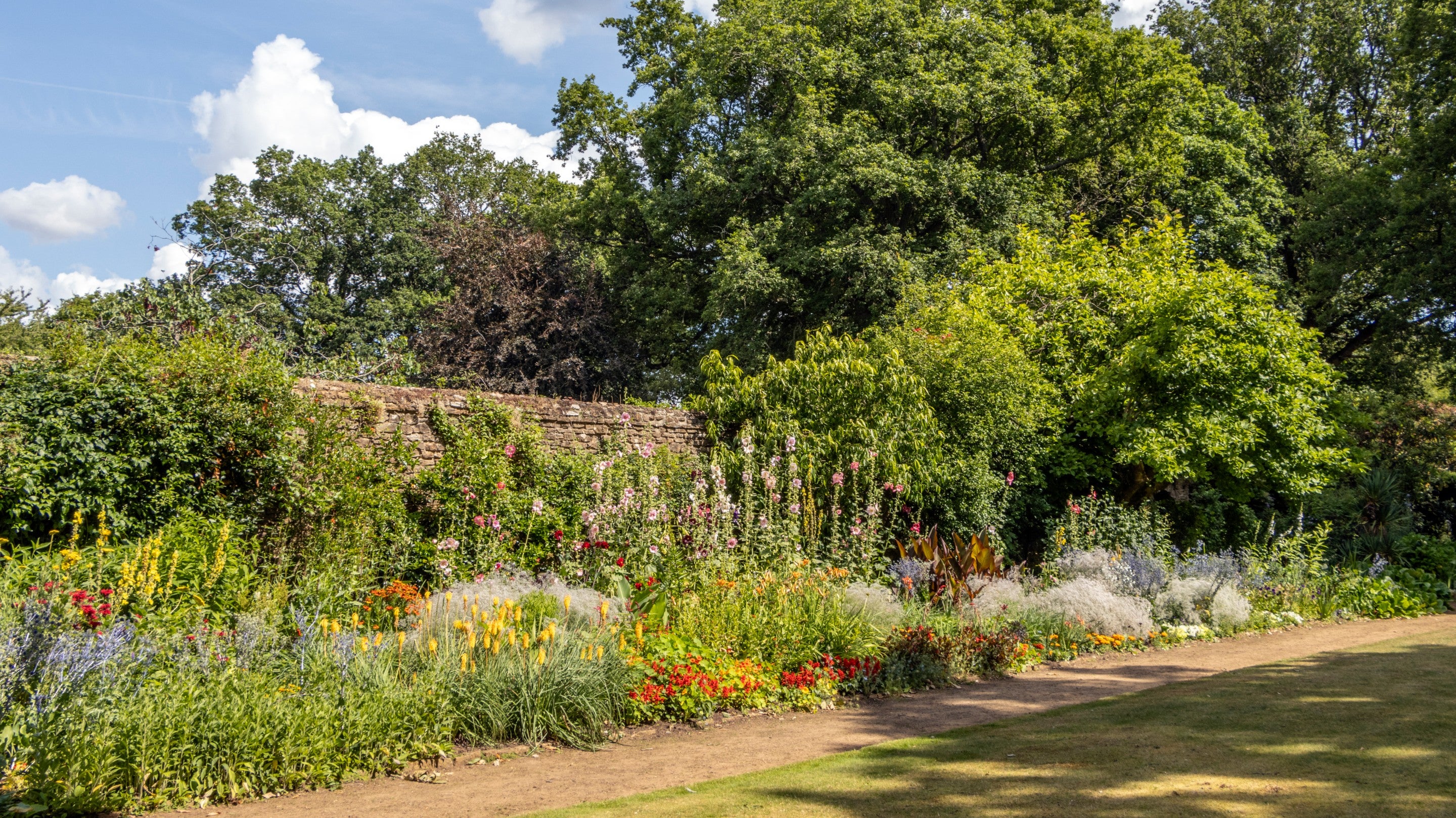 A view of the main flower border at Munstead Wood. Red and yellow flowers grow in front of climbing plants on a stone wall, overshadowed by trees in the background