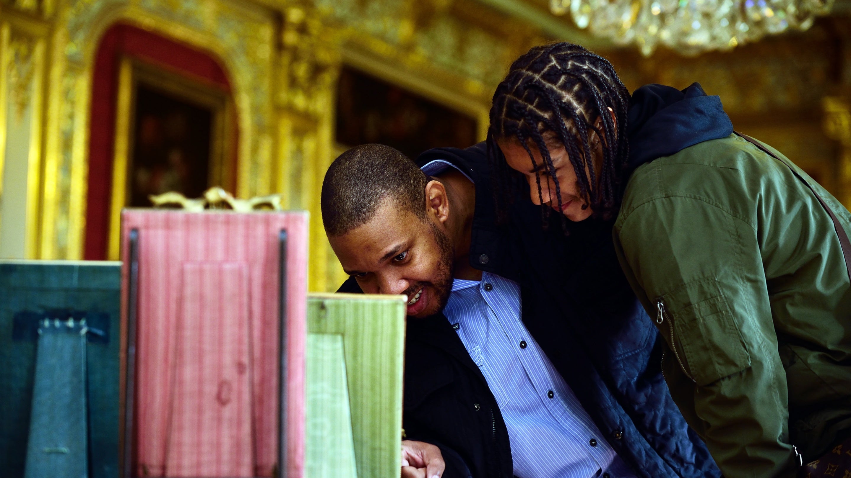 Two people look at a green coloured photo frame with gold coloured walls and chandelier in the background
