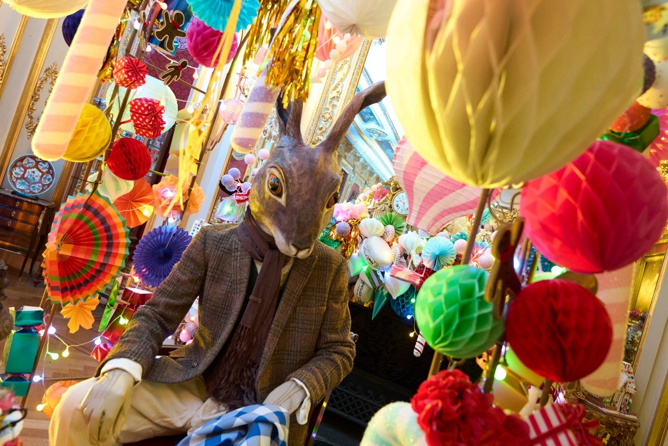 Alice's Christmas in Wonderland at Polesden Lacey 2025: a large, human-sized hare mannequin, dressed in a brown jacket and scarf, sits at a tea table with brightly coloured Christmas decorations behind him
