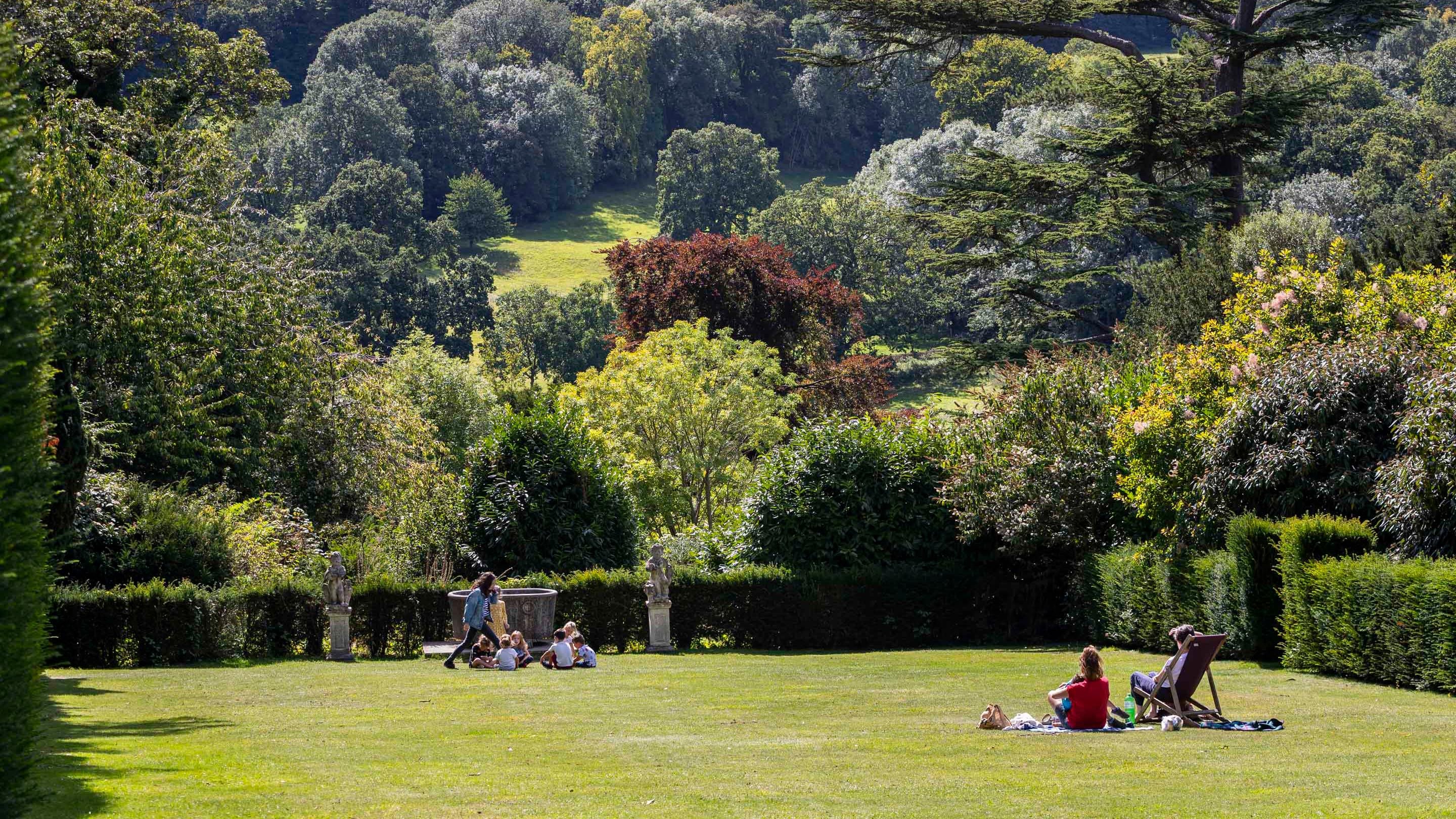 Visitors in the garden at Polesden Lacey on a summer's day. A couple sit having a picnic on the lawn and a group of children play a game further down the lawn. The lawn is edged by a hedge and beyond the parkland filled with mature trees can be seen stretching up the hill into the distance.