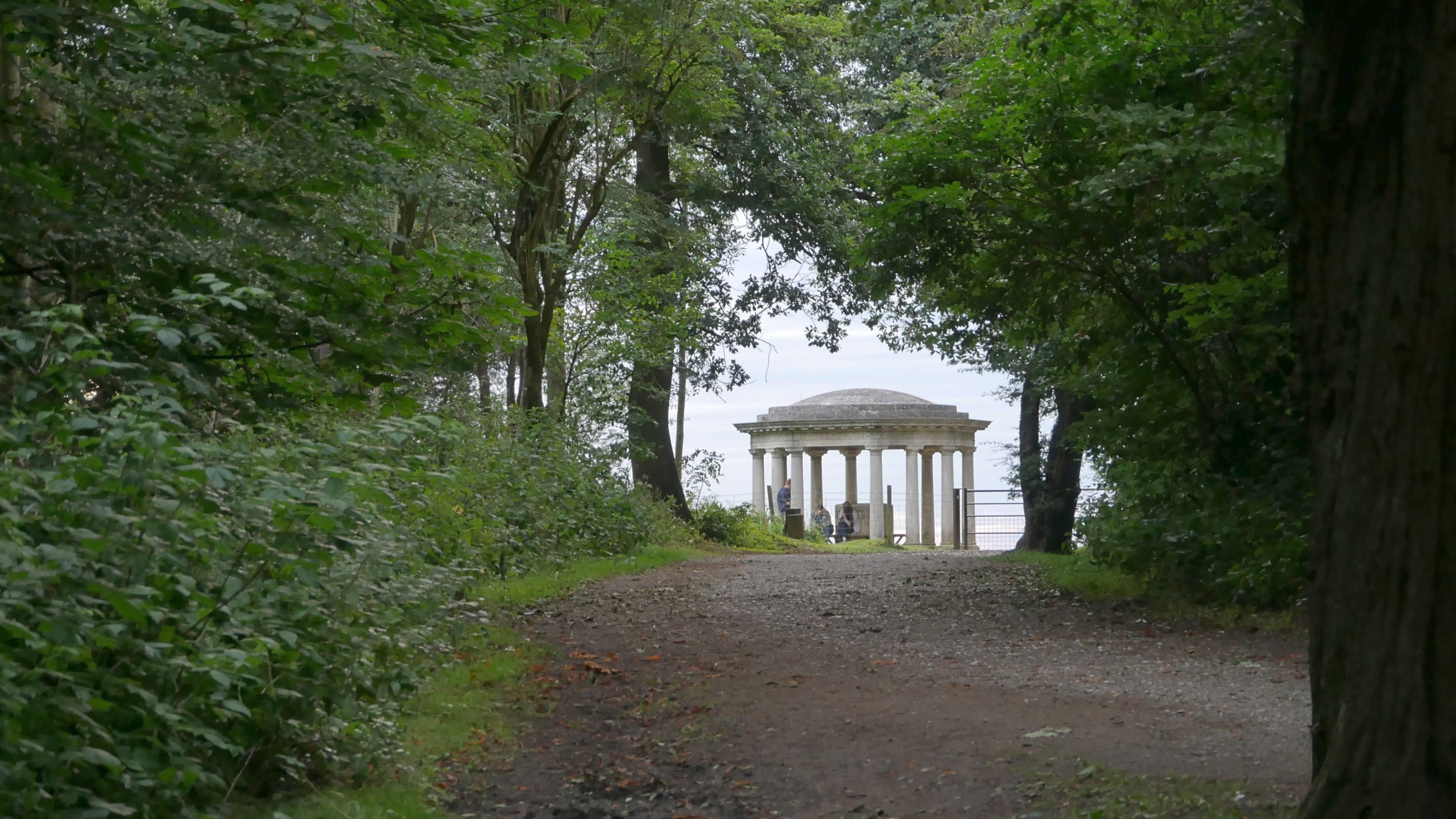 A view through woods to a tall, white, circular Inglis memorial structure with a domed roof and column details at Reigate Hill and Gatton Park in Surrey