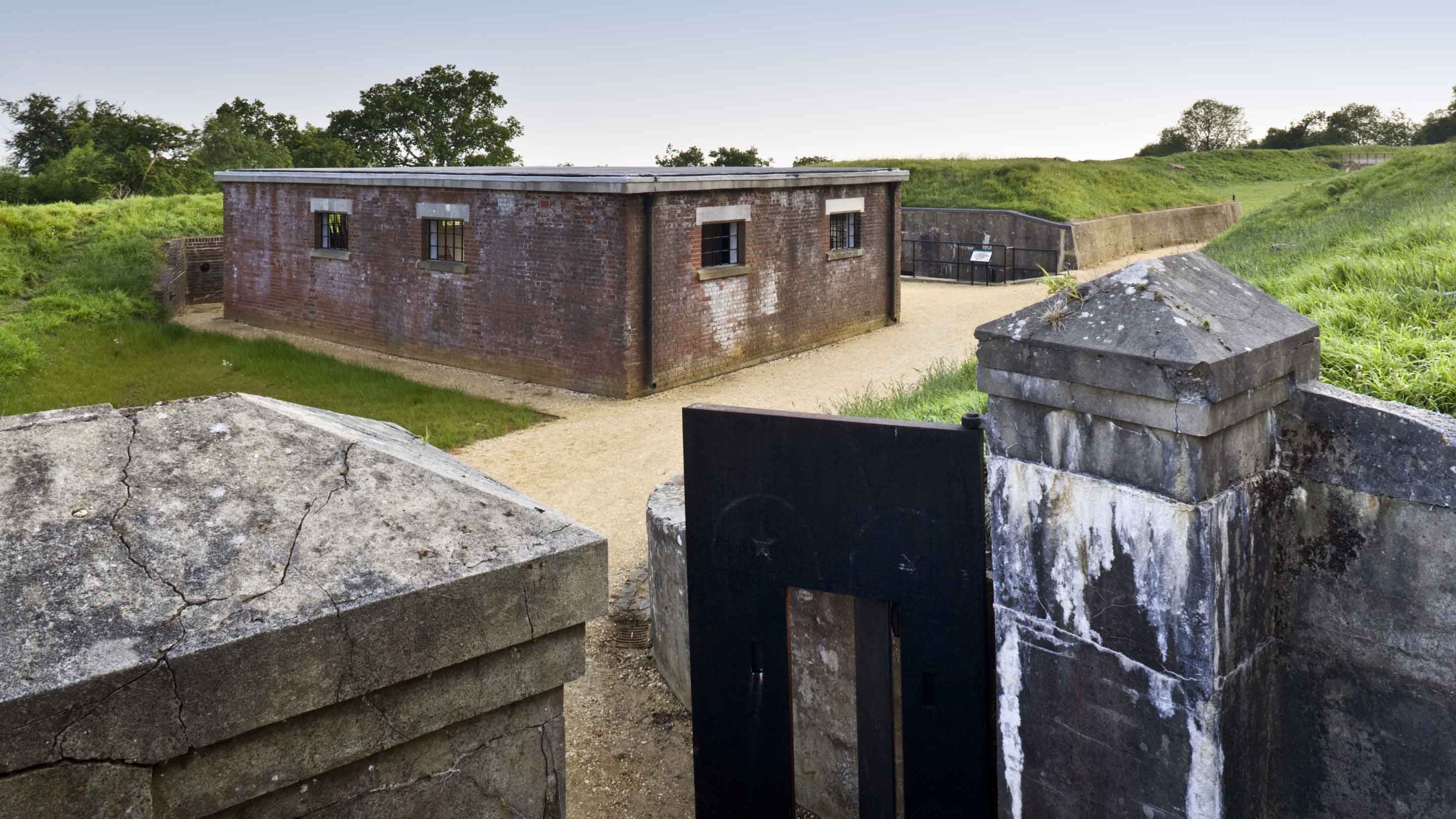 The Tool Store seen from the entrance gates at Reigate Fort, Surrey. A short square building surrounded by grass, with gates in the foreground.