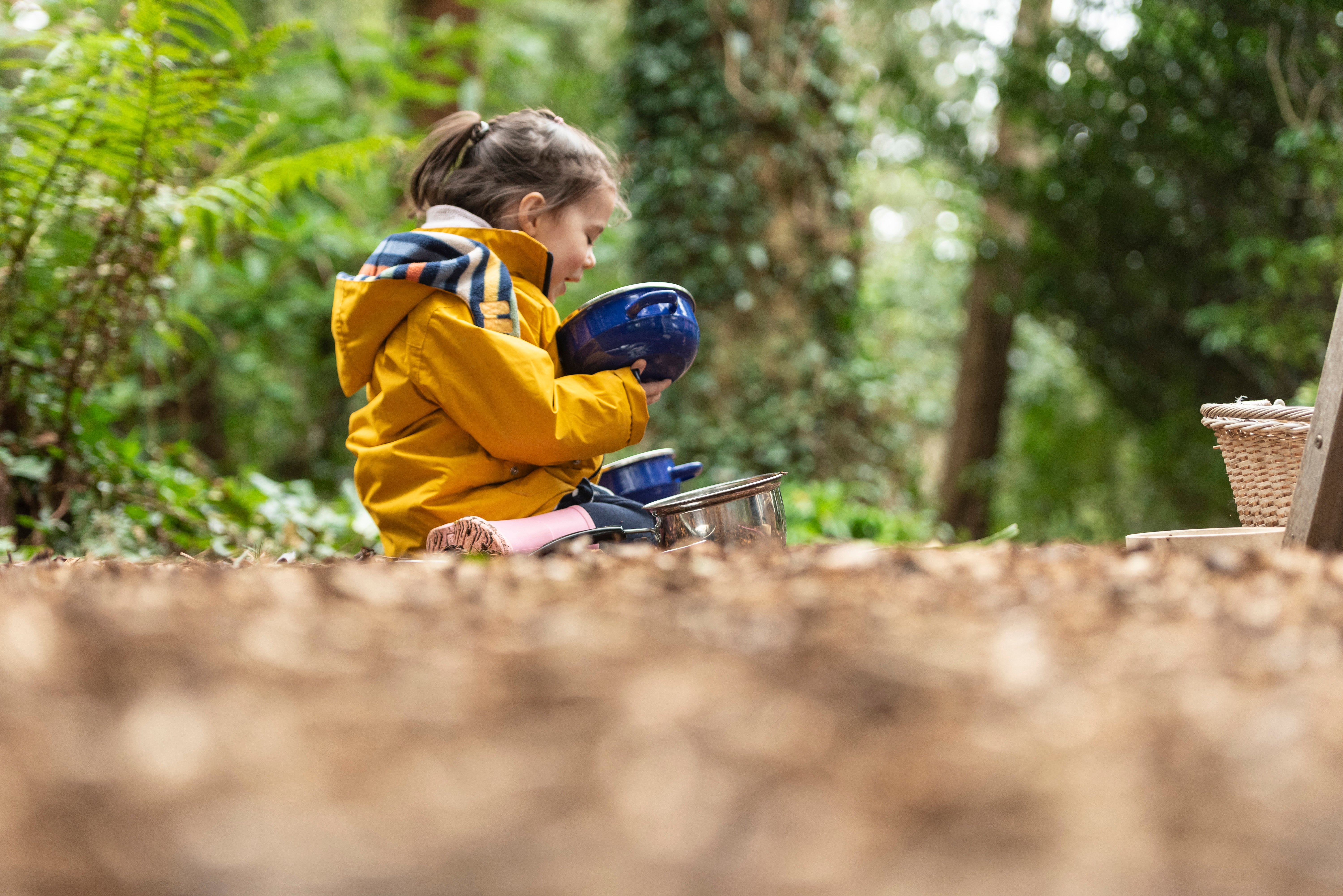 Young child in a yellow raincoat sitting on the forest floor among leaves playing with a saucepan