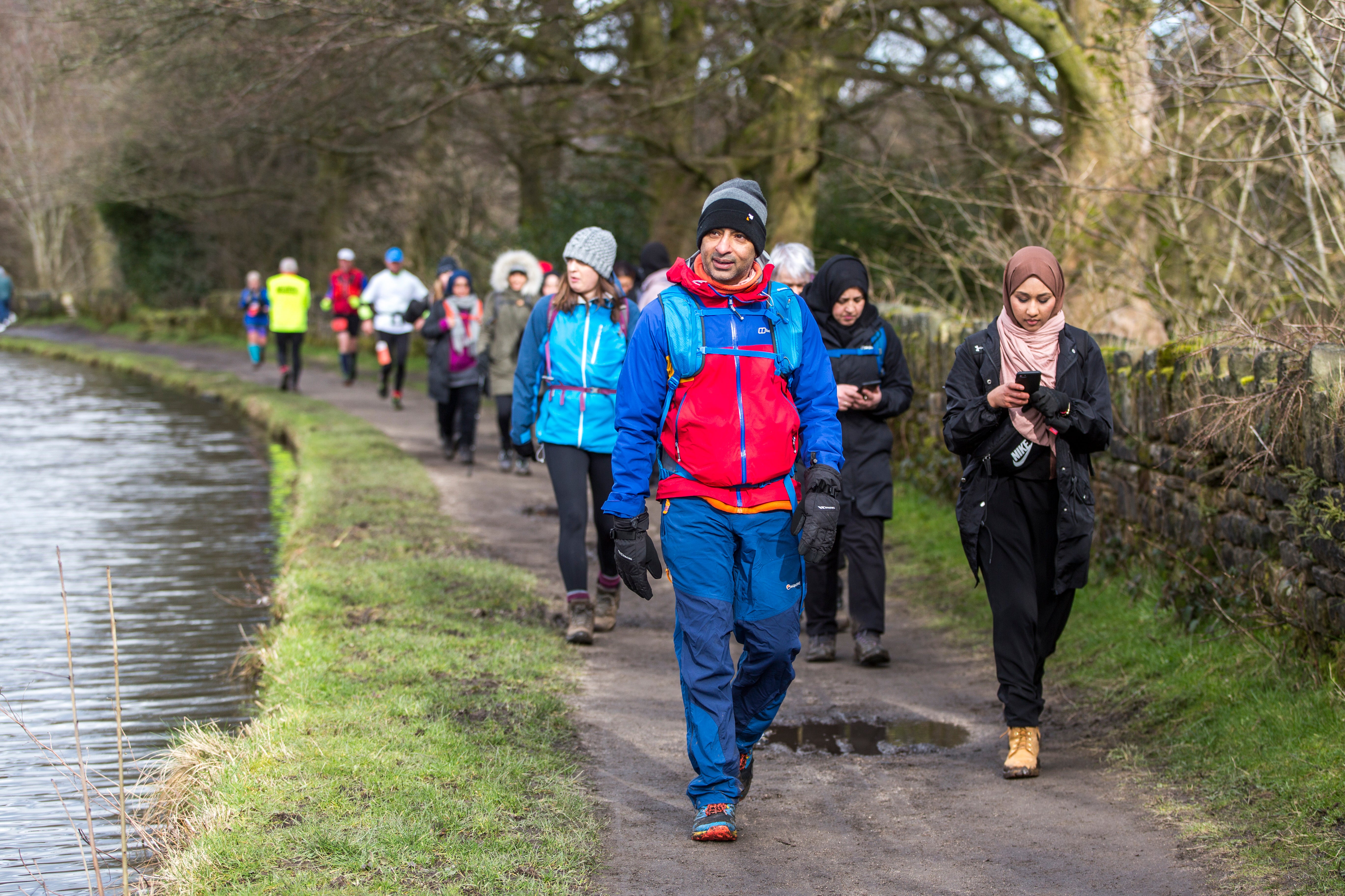 A group of people in waterproof coats and woolly hats, walking on a path beside a waterway.