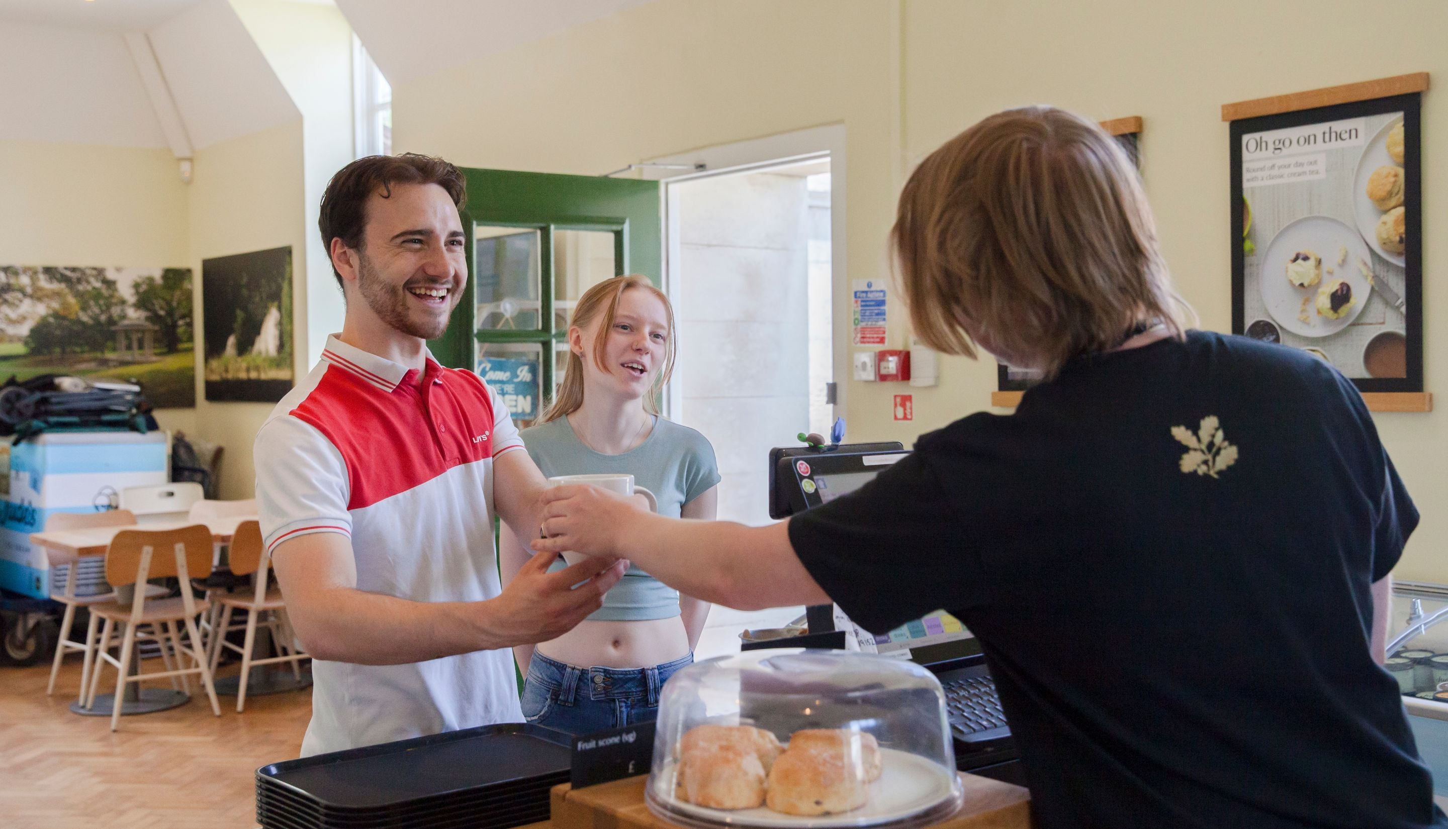 A café counter where a staff member is handing a drink to a customer. On the counter, there is a glass dome with scones inside and a payment terminal. In the background, there are tables, chairs, and posters on the wall. The setting is bright and casual.