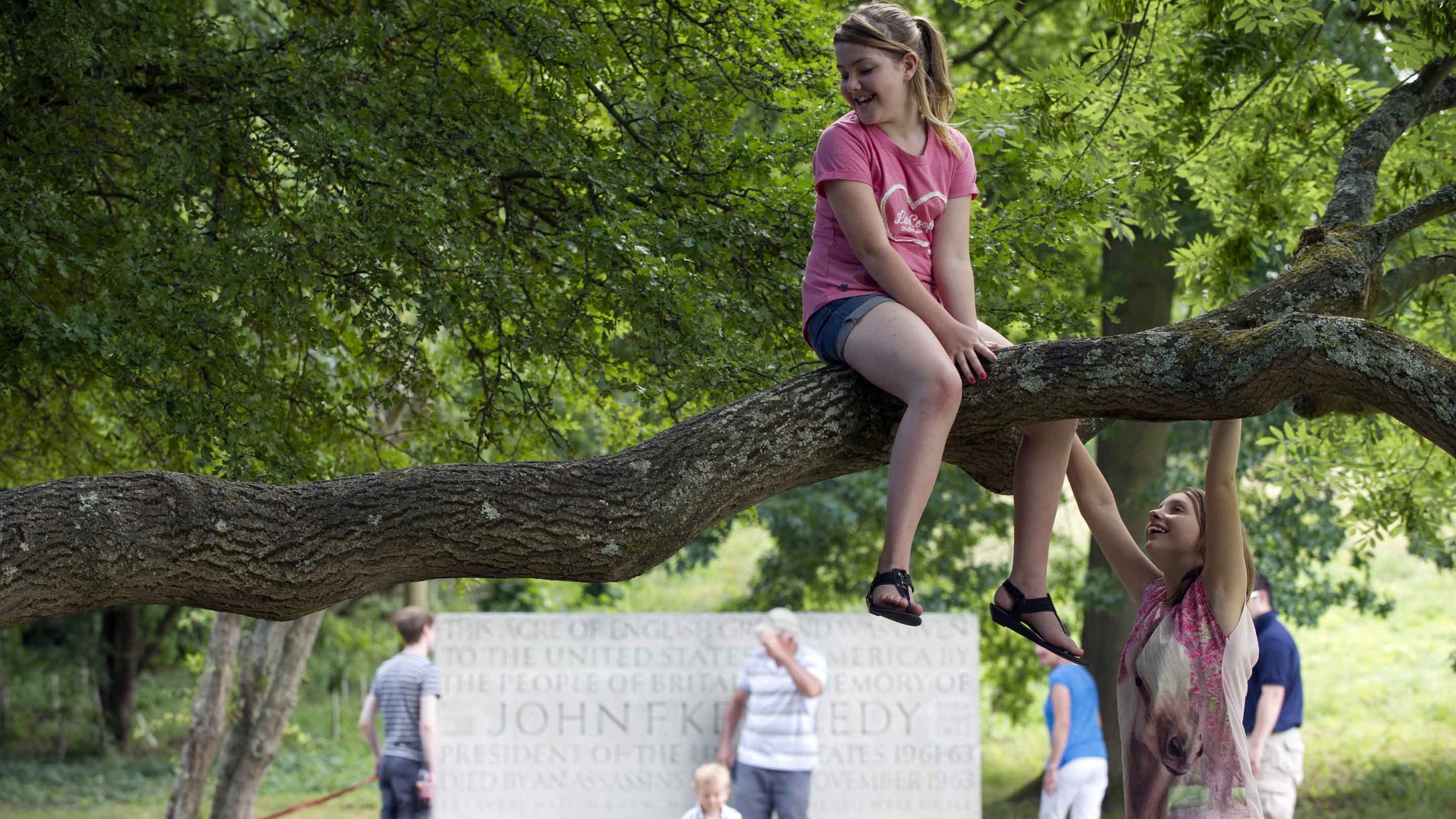 Visitors standing in front of the Kennedy Memorial, Runnymede, Surrey, with a girl sitting on a tree branch in the foreground, with another girl nearby. This memorial to assassinated US president John F Kennedy (1917-1963) was designed by Sir Geoffrey Jellicoe. The words inscribed on the 7-ton block of pale cream-coloured Portland stone are taken from President Kennedy's inaugural address in 1961