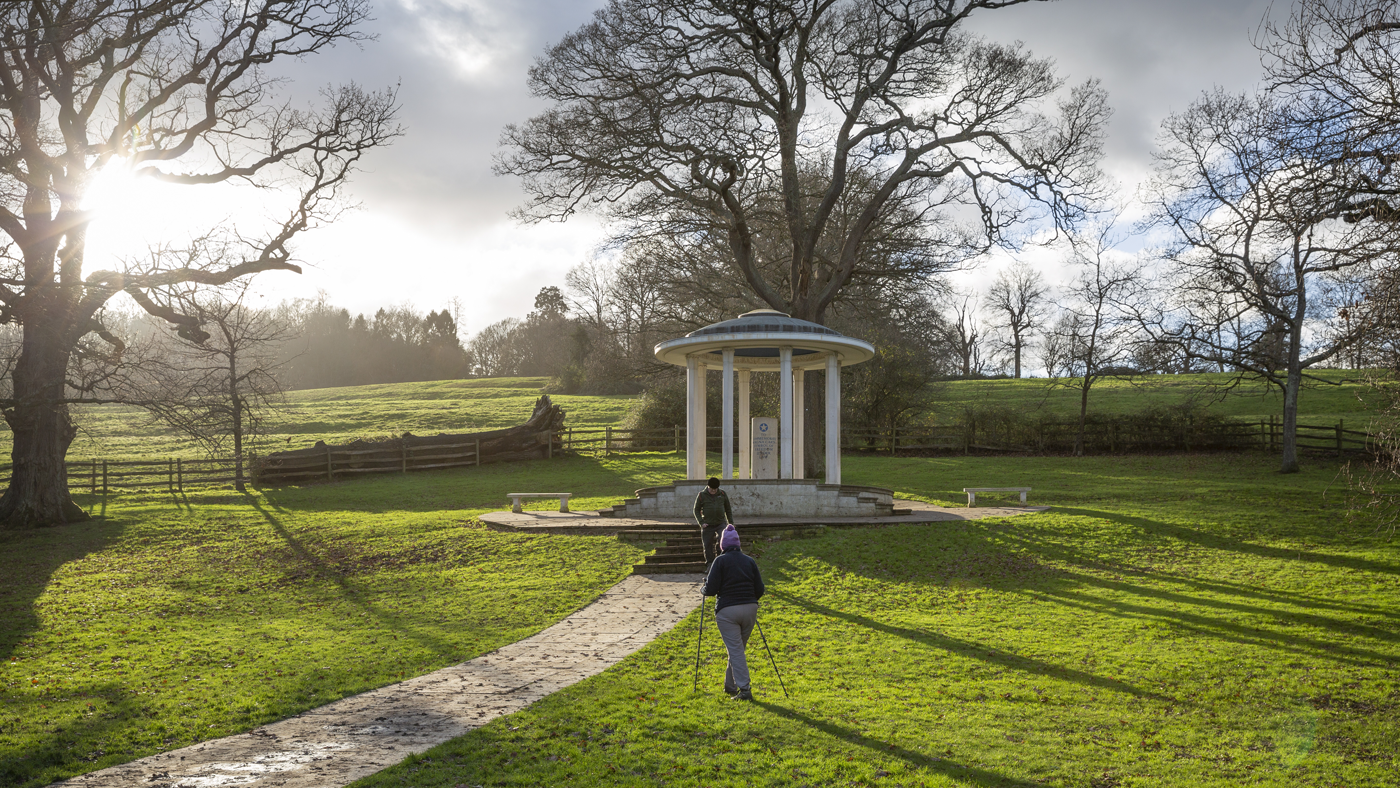 A couple walking towards the Magna Carta Memorial - a domed structure- they're walking with rambling poles through the slightly muddy looking field with bare wintry trees in the background and low winter sun rays.