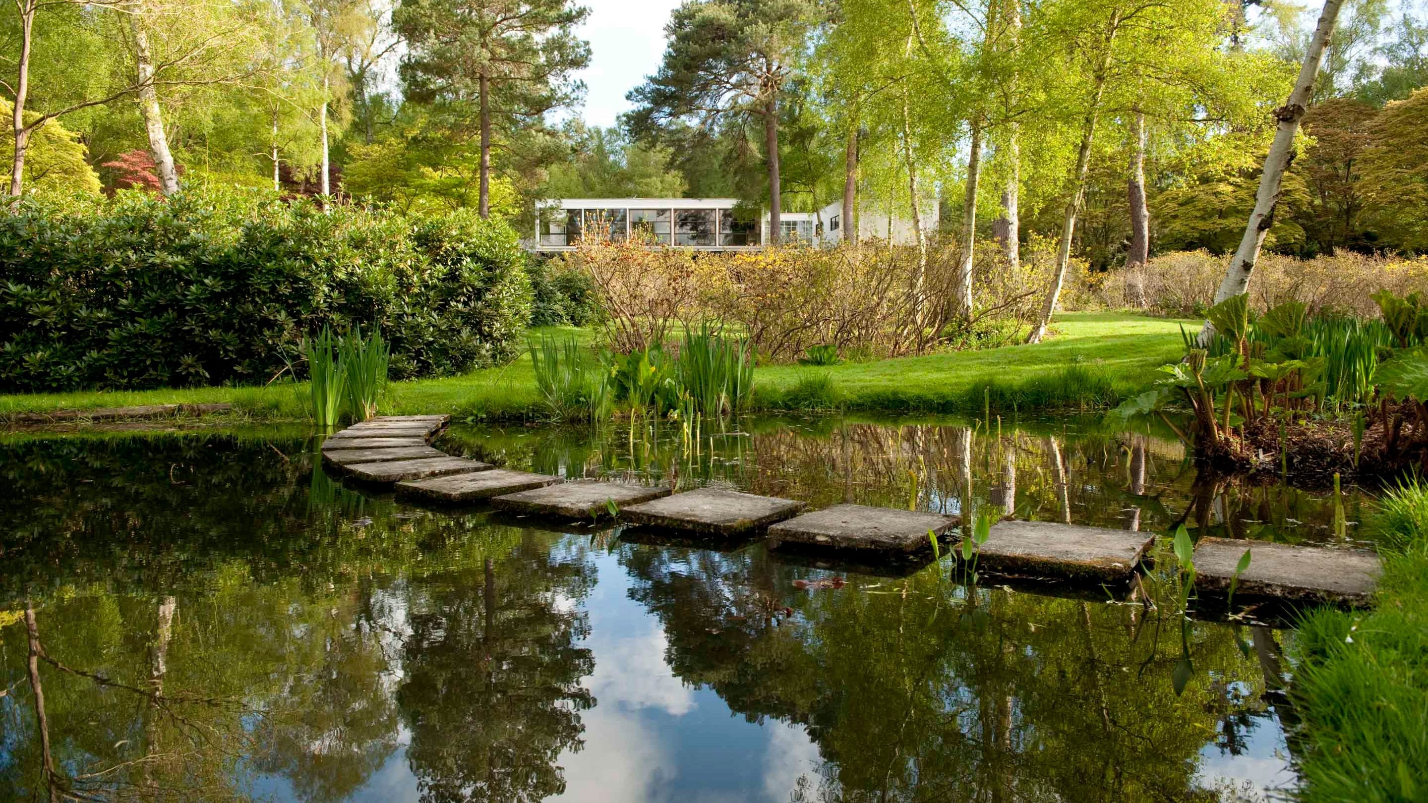 A view of the garden at The Homewood, Surrey. In the foreground is a pond with stepping stones across it, trees and shrubbery surround the garden and in the distance the exterior of the house can be seen.