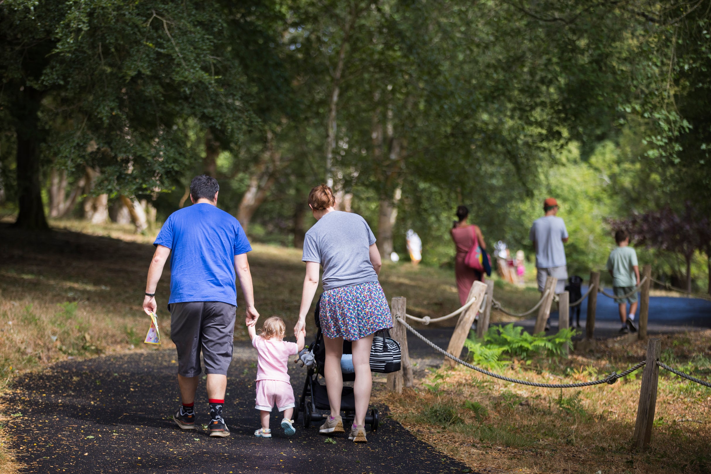 Mother and father holding their child's hands walking along a tarmacked pathway at Winkworth Arboretum. Mum is also pushing a pram. There are other people walking in the background.