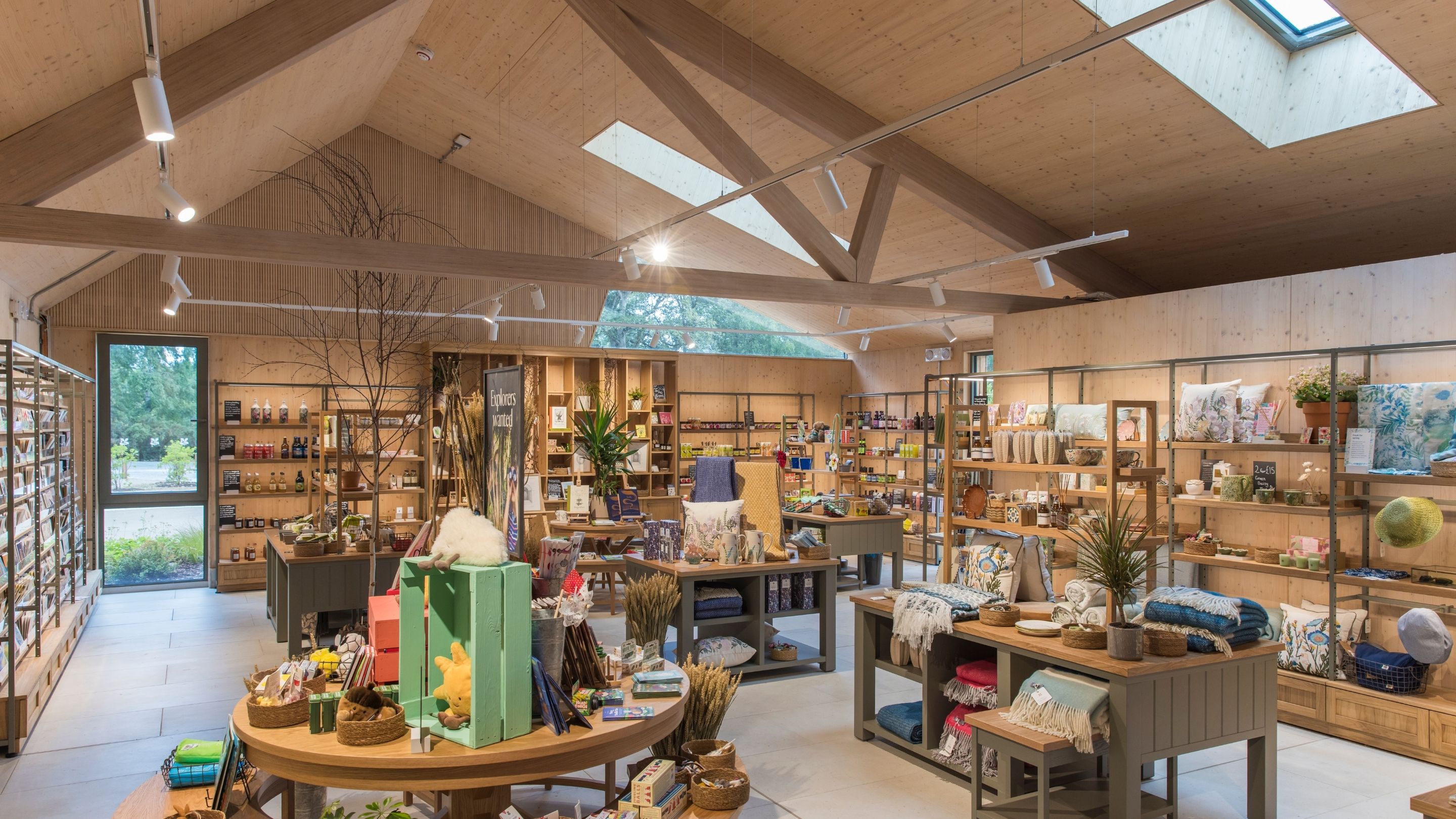 Retail shop interior with exposed timber beams, skylights, and wooden shelving displaying gifts, homewares, books, plants, and local products. Central display tables create an open, welcoming shopping space.