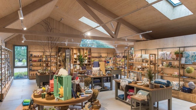 Retail shop interior with exposed timber beams, skylights, and wooden shelving displaying gifts, homewares, books, plants, and local products. Central display tables create an open, welcoming shopping space.