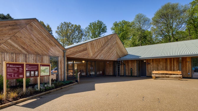 The courtyard outside the entrance to the visitor welcome building. To the left are some information points, there is a wooden bench with toilet facilities behind on the right.