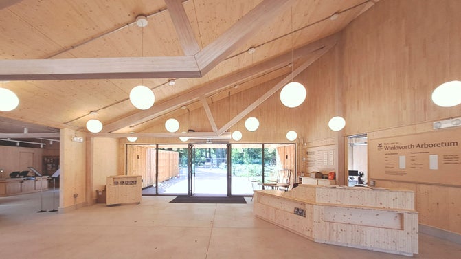 A bright and area foyer space clad in a light wooden material with globe lights hanging from the building.  There is a large reception desk with the right of the area with signage displaying pricing information.