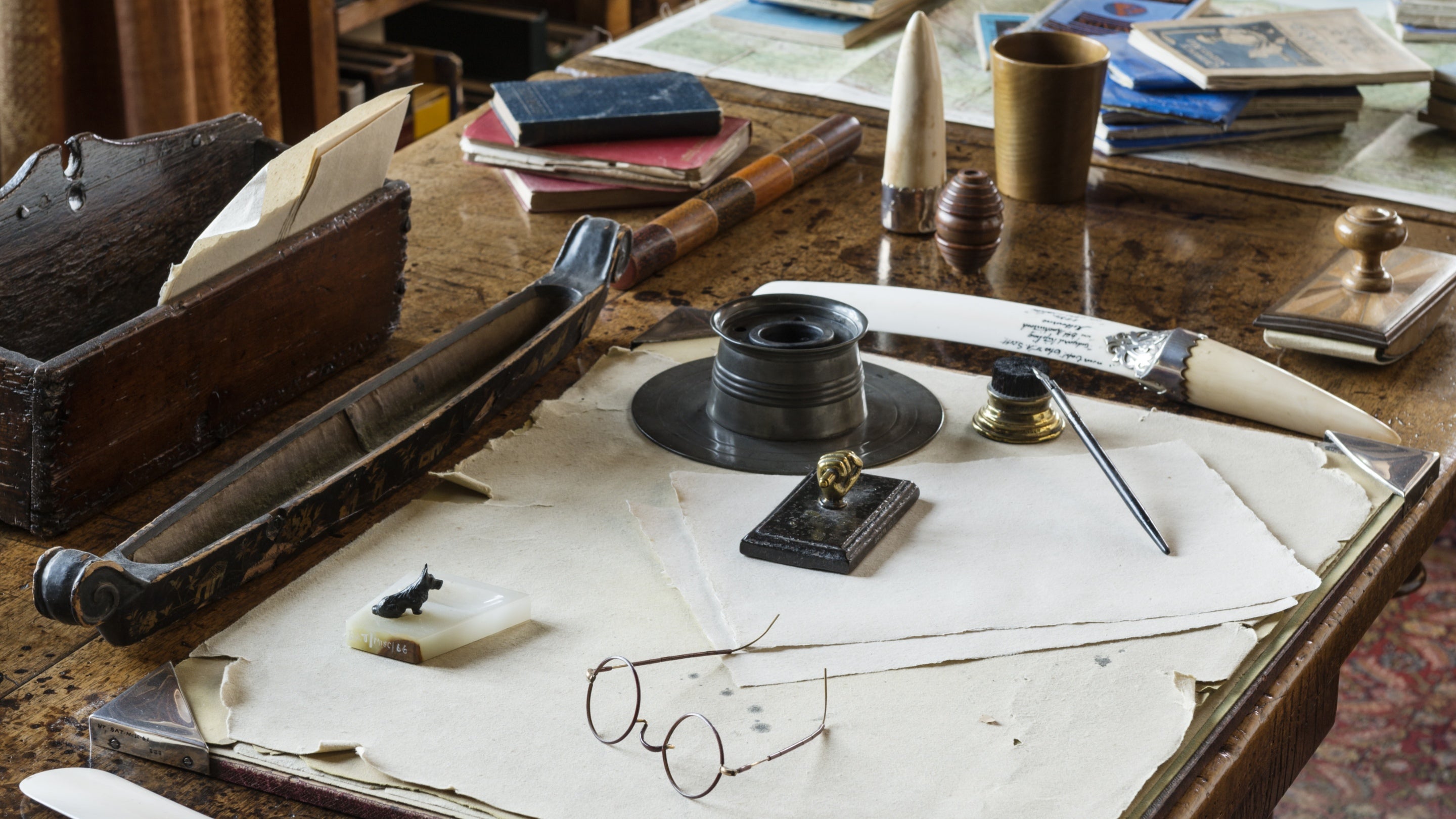A close up image of Rudyard Kipling's wooden desk inside Bateman's with his thin wire framed glasses, ink well and various ornaments set out on its surface