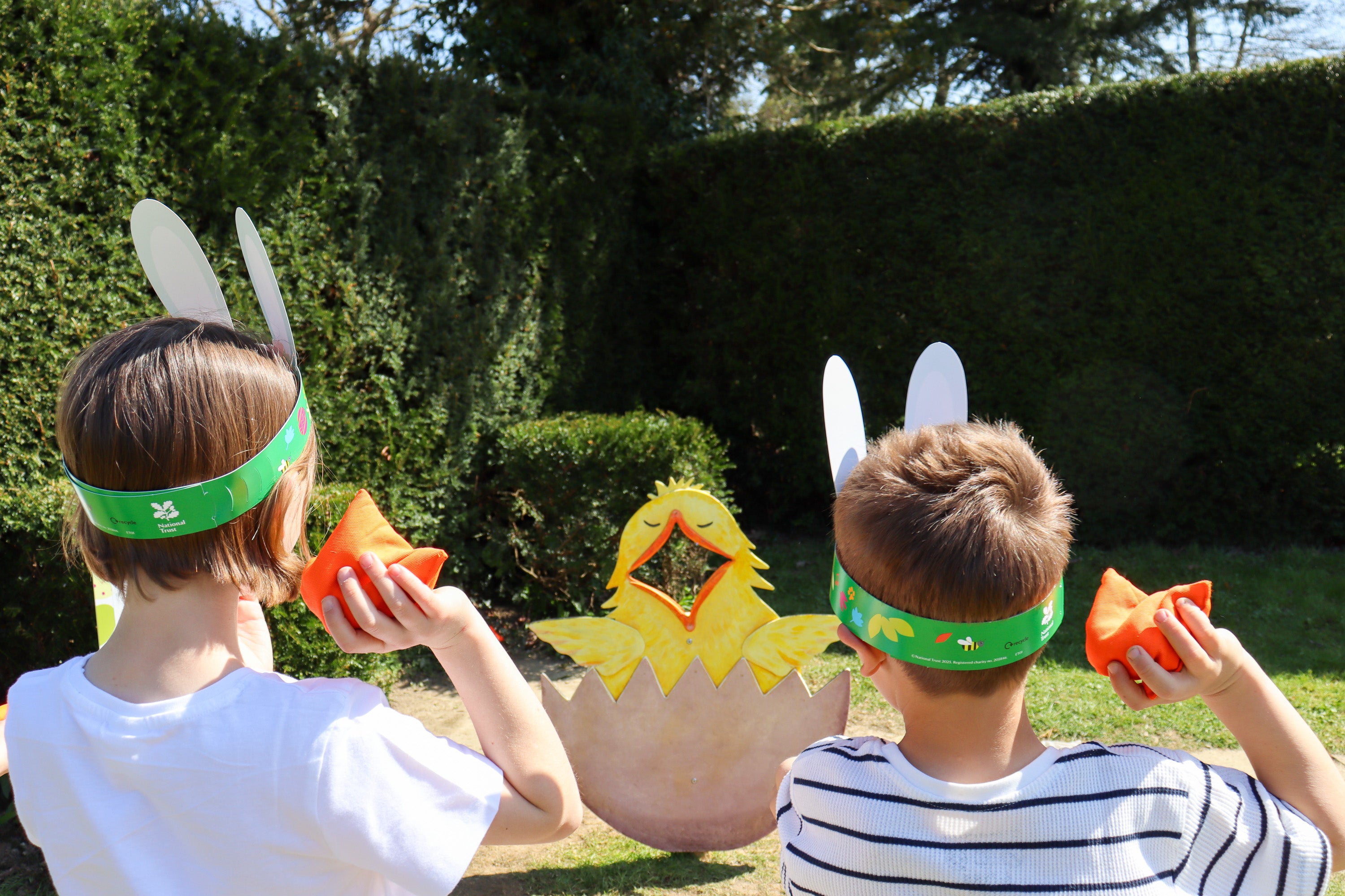 Two people with their backs to the camera holding an orange bean bag ready to throw at a wooden chick with it's mouth open.