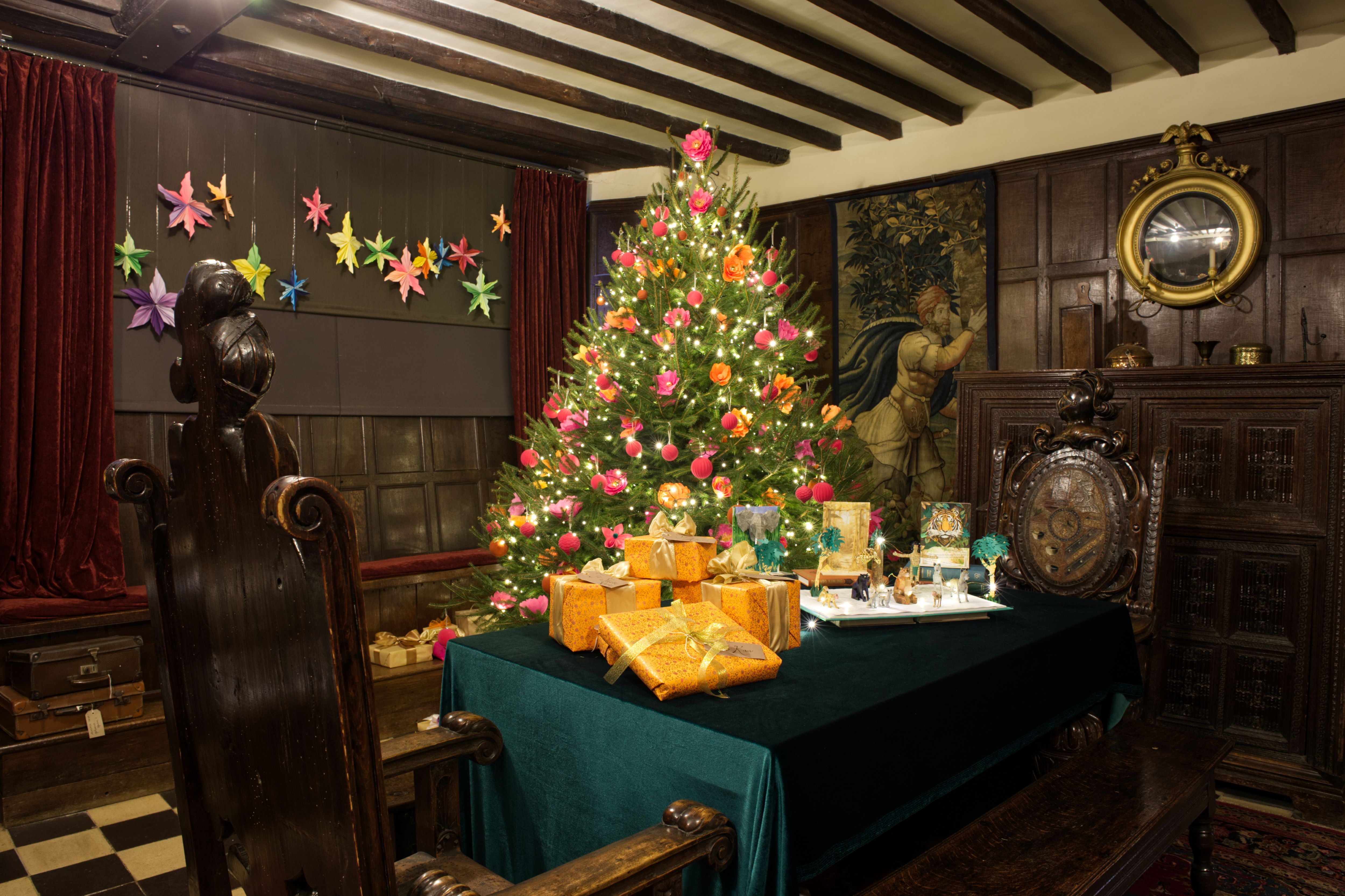 Historic room decorated for Christmas with a glowing tree, colourful ornaments, and wrapped gifts on a green-covered table.