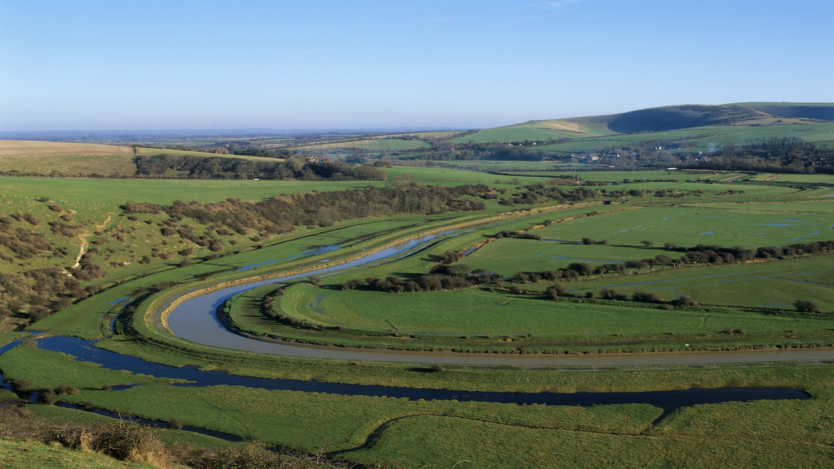 A wide, sweeping view of a green river valley with a winding river cutting through open fields. Low, gently rolling hills stretch into the distance under a clear blue sky, with patches of shrubs and hedgerows scattered across the landscape