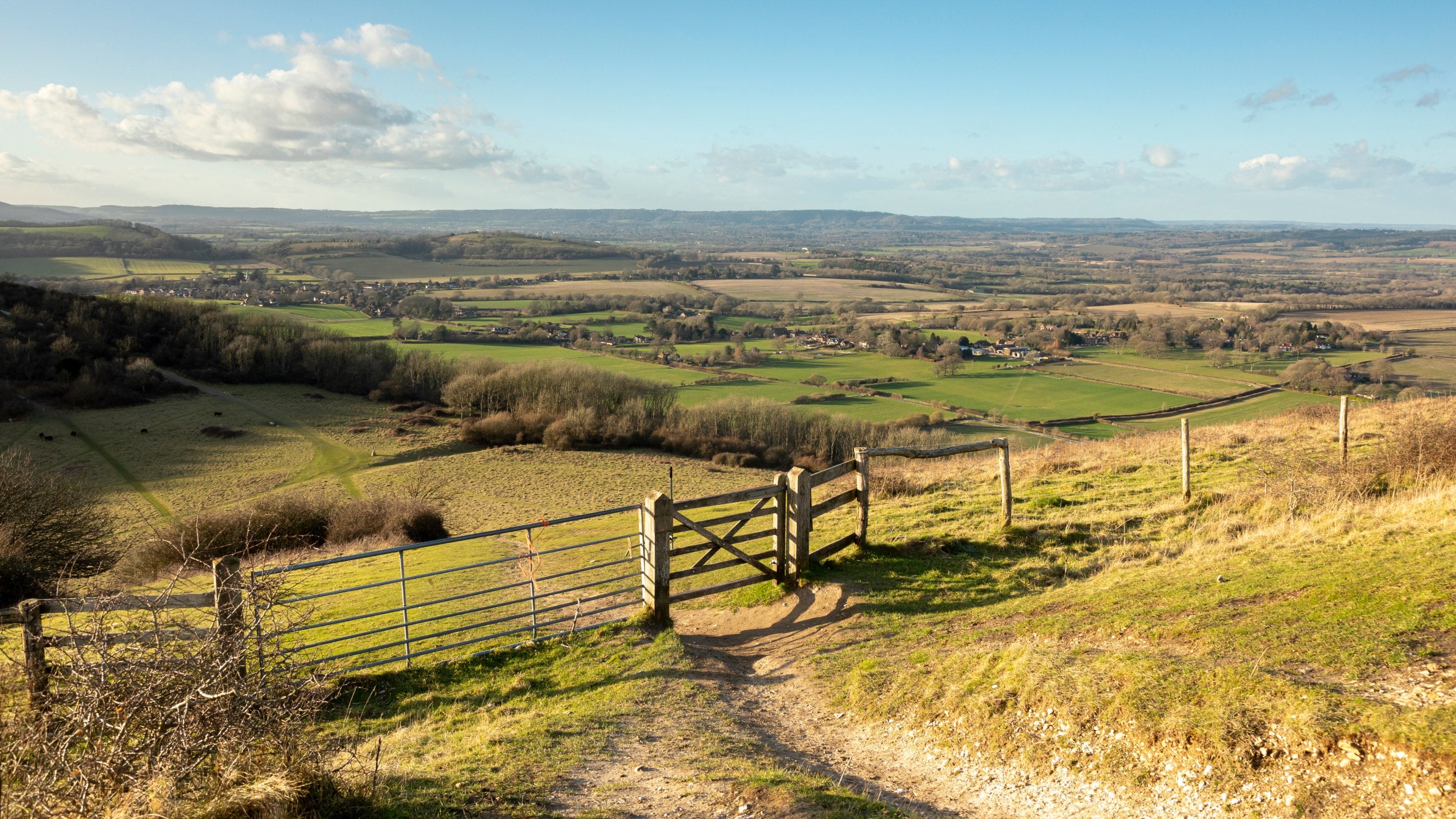 View from Harting Down, West Sussex, where a path is crossed by a wooden gate, with views downhill to a patchwork of fields and woodland
