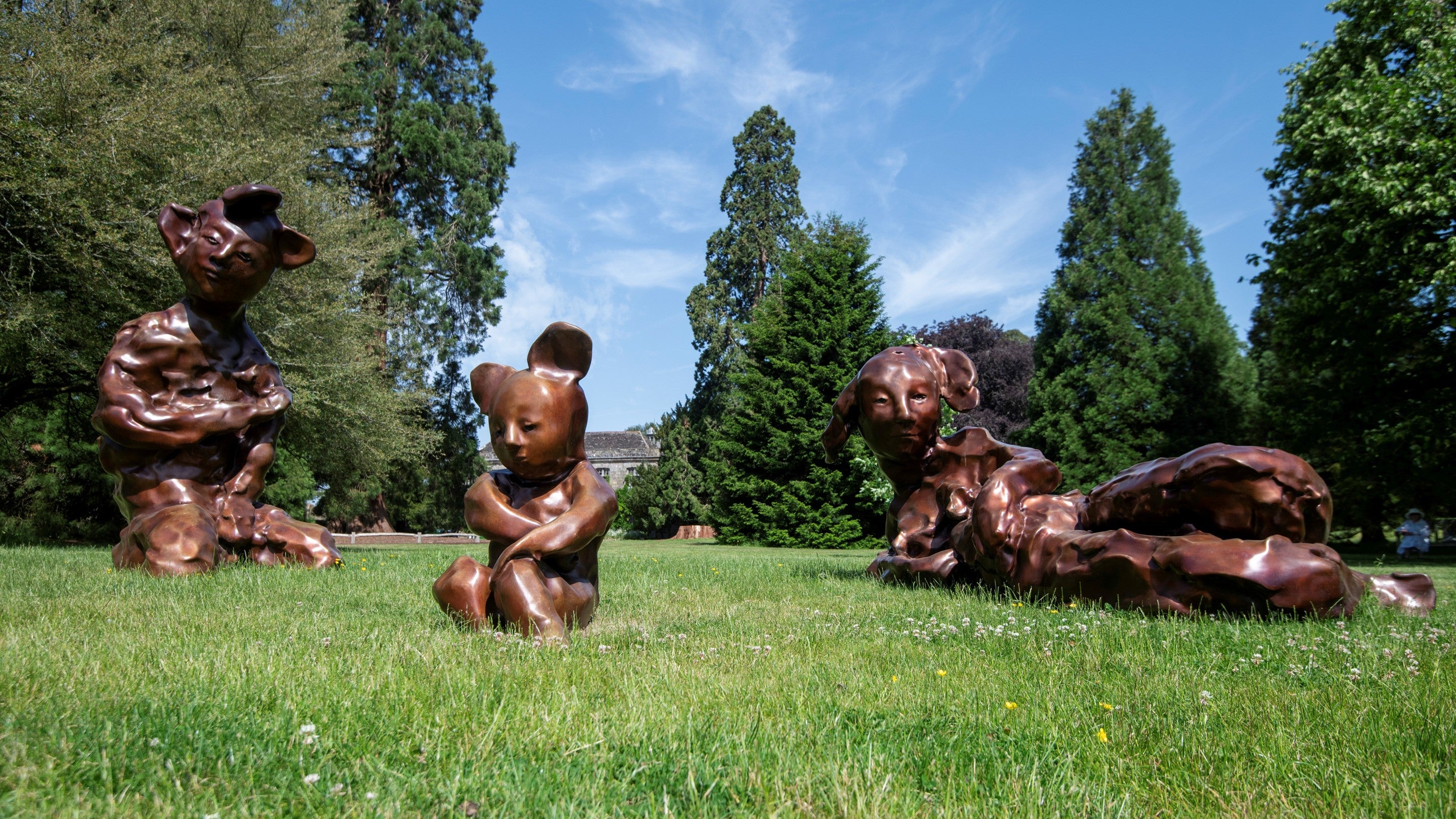 Three large bronze sculptures depicting animal-human hybrid figures sit on a lawn at Wakehurst, surrounded by towering trees beneath a blue summer sky.