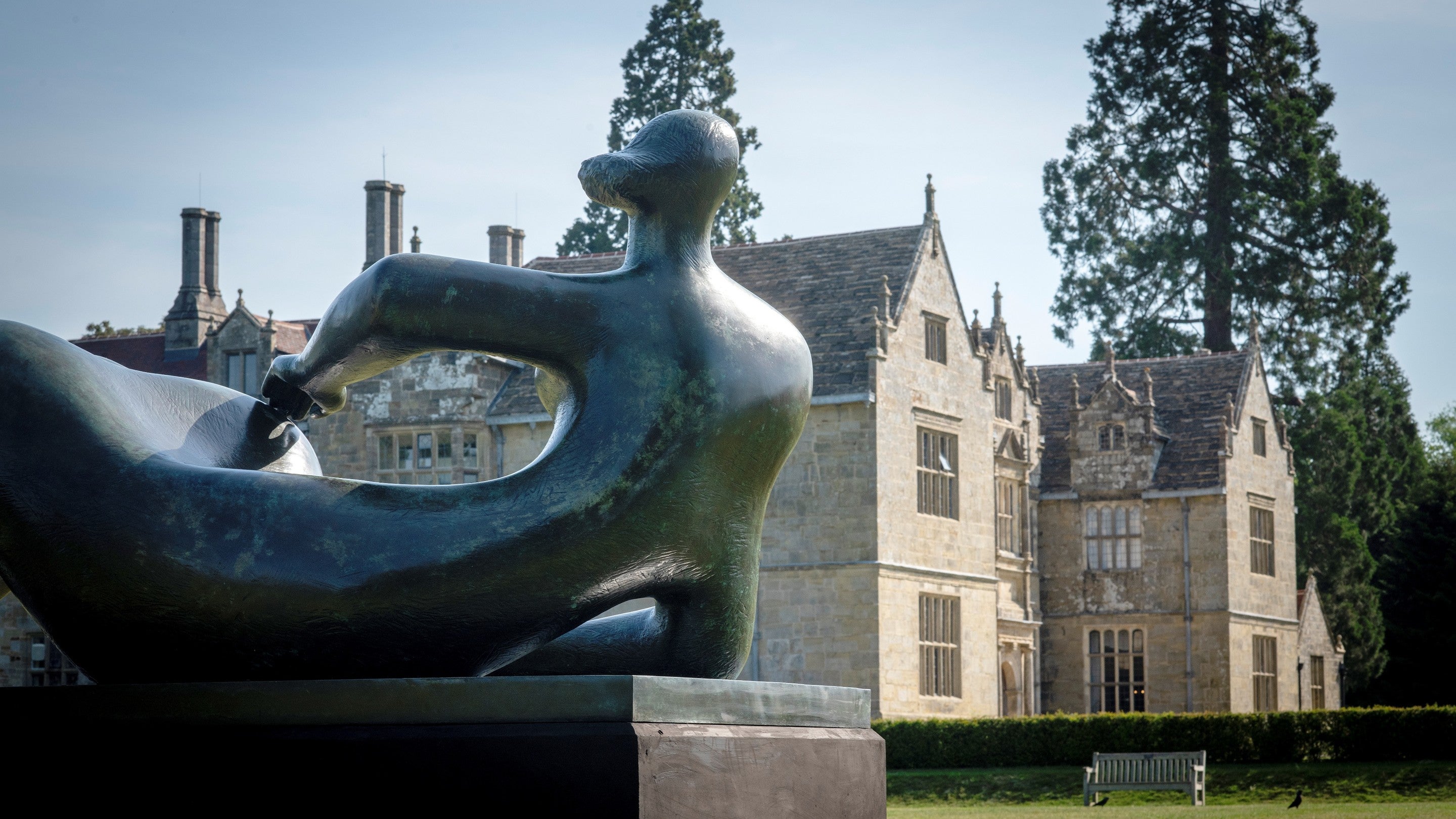 Henry Moore’s bronze sculpture Reclining Figure stands on a plinth in front of the historic Wakehurst mansion, with mature trees and lawns in the background.