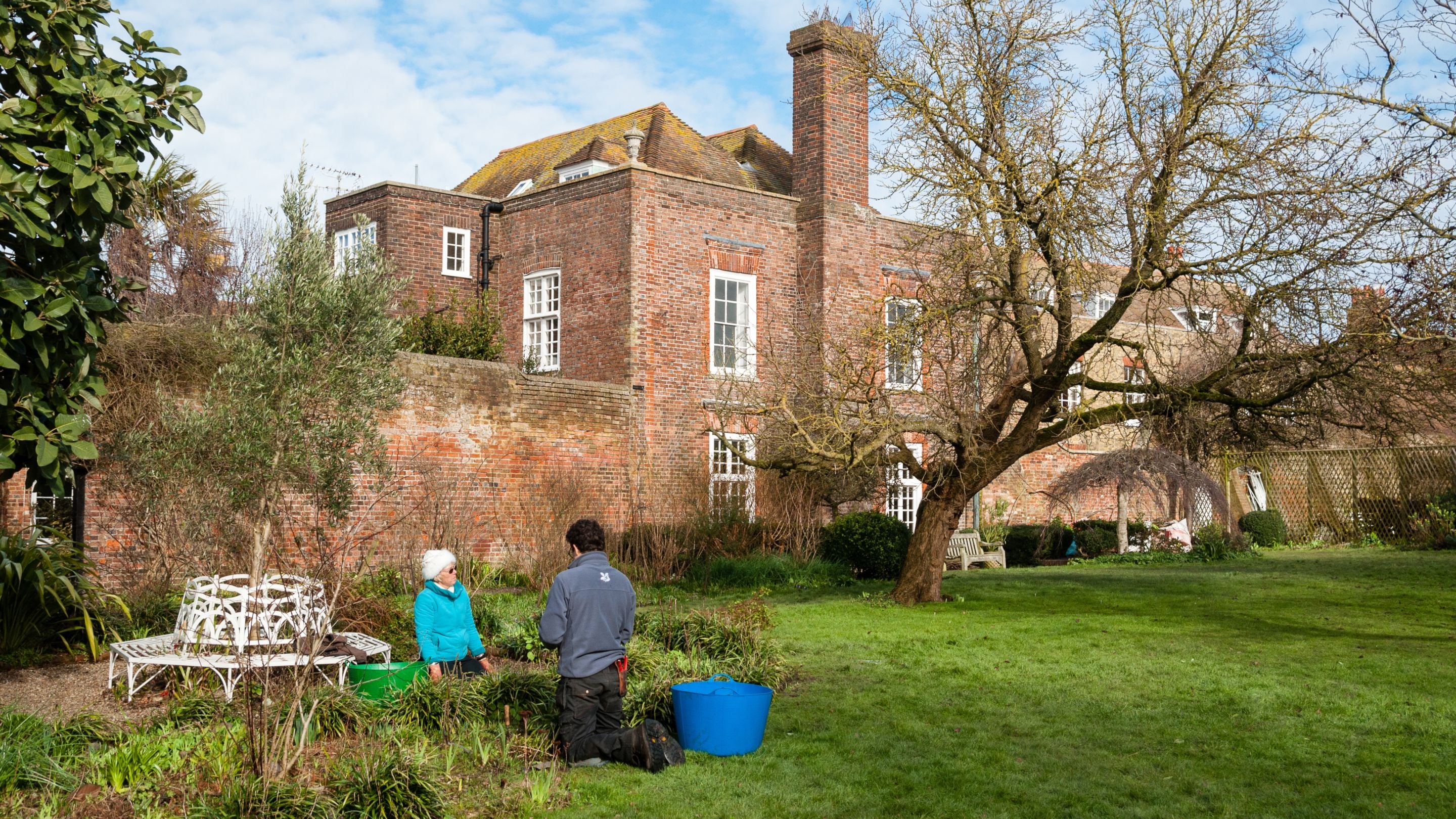 Two garden volunteers kneeling on the grass at Lamb House, East Sussex, near a circular white wrought-iron bench with the house in the background