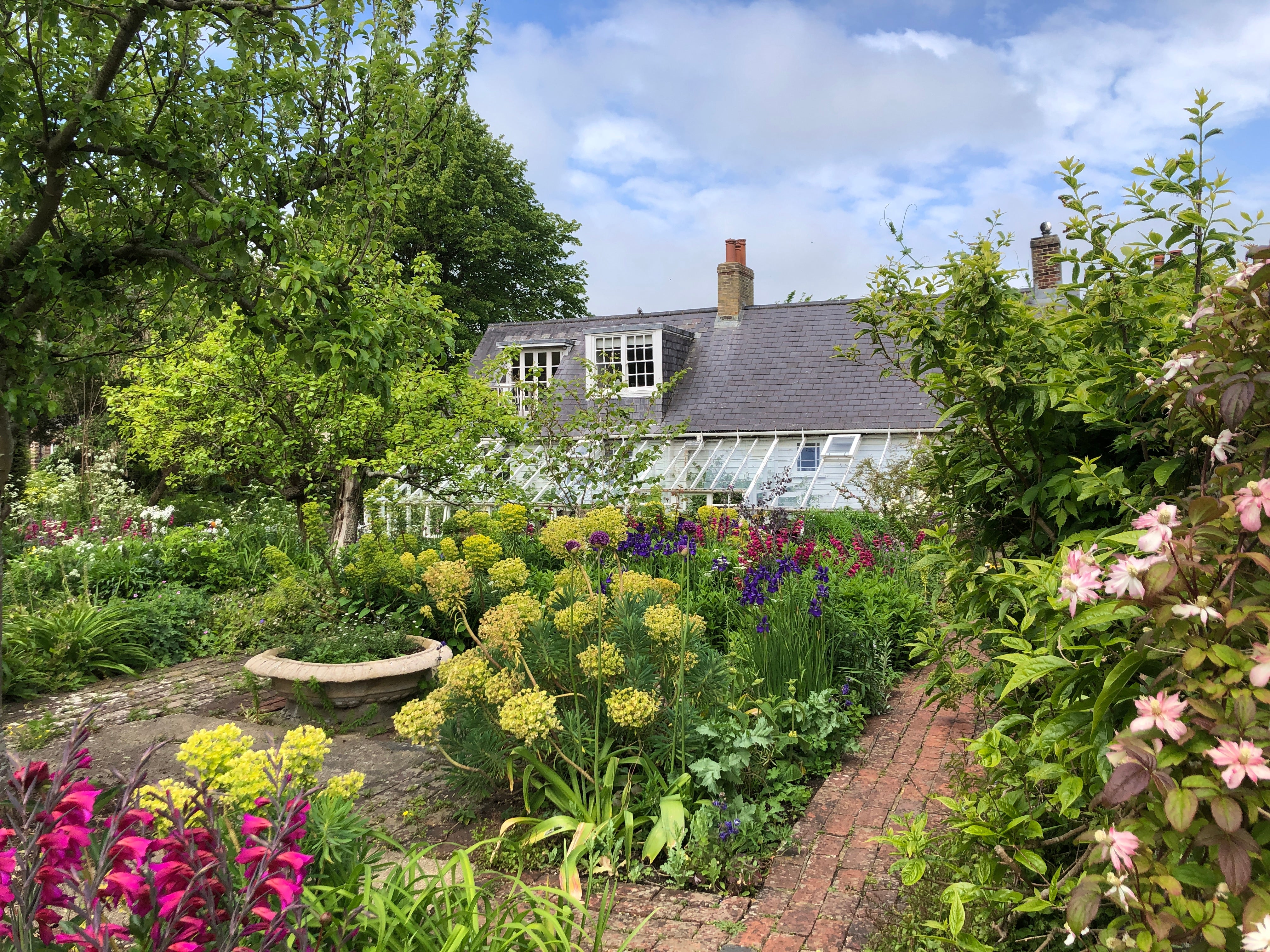 Monk's House and conservatory viewed through the colourful florals in the cottage garden. There is a blue sky with white clouds