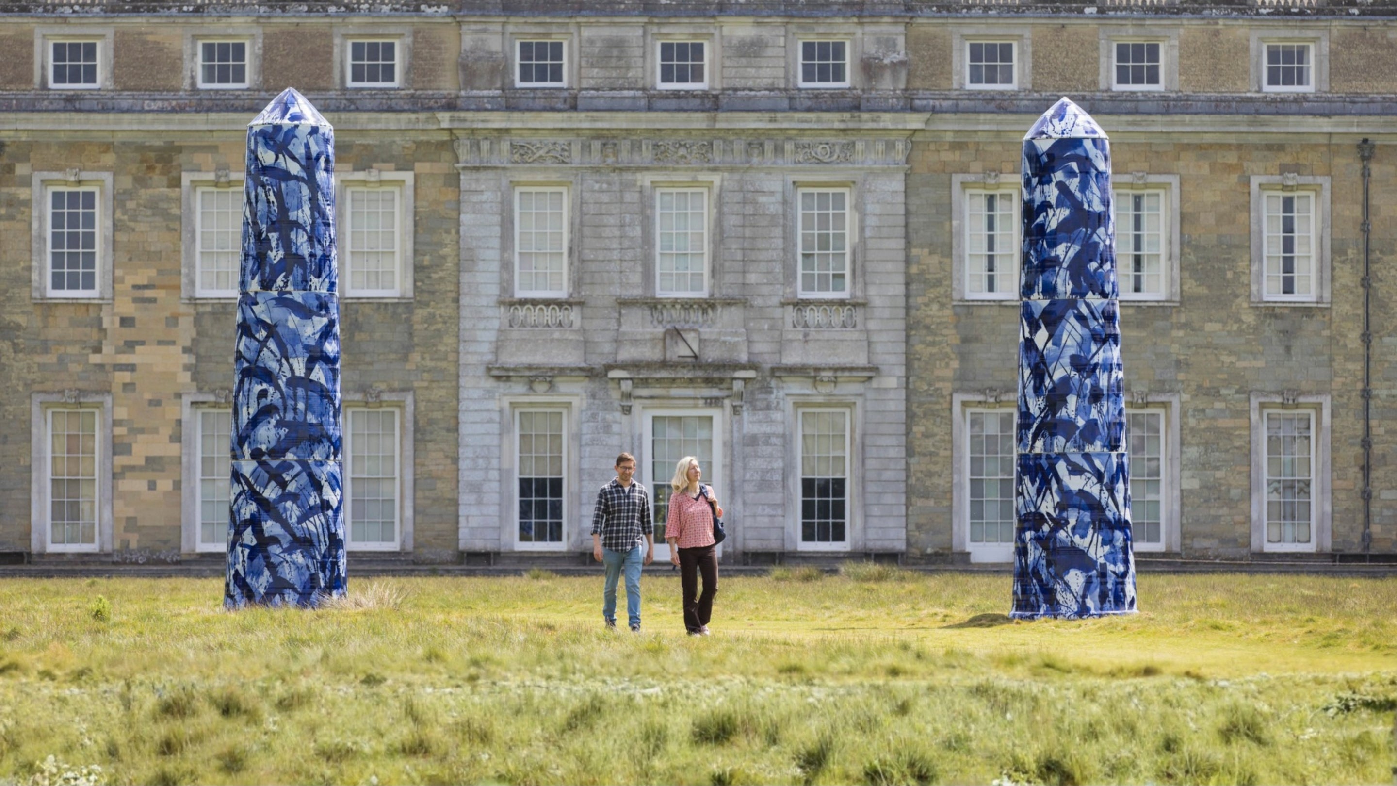 Two visitors walking past giant blue and white ceramic monoliths from the Expressions in Blue exhibition, in Petworth House's parkland with house in background.